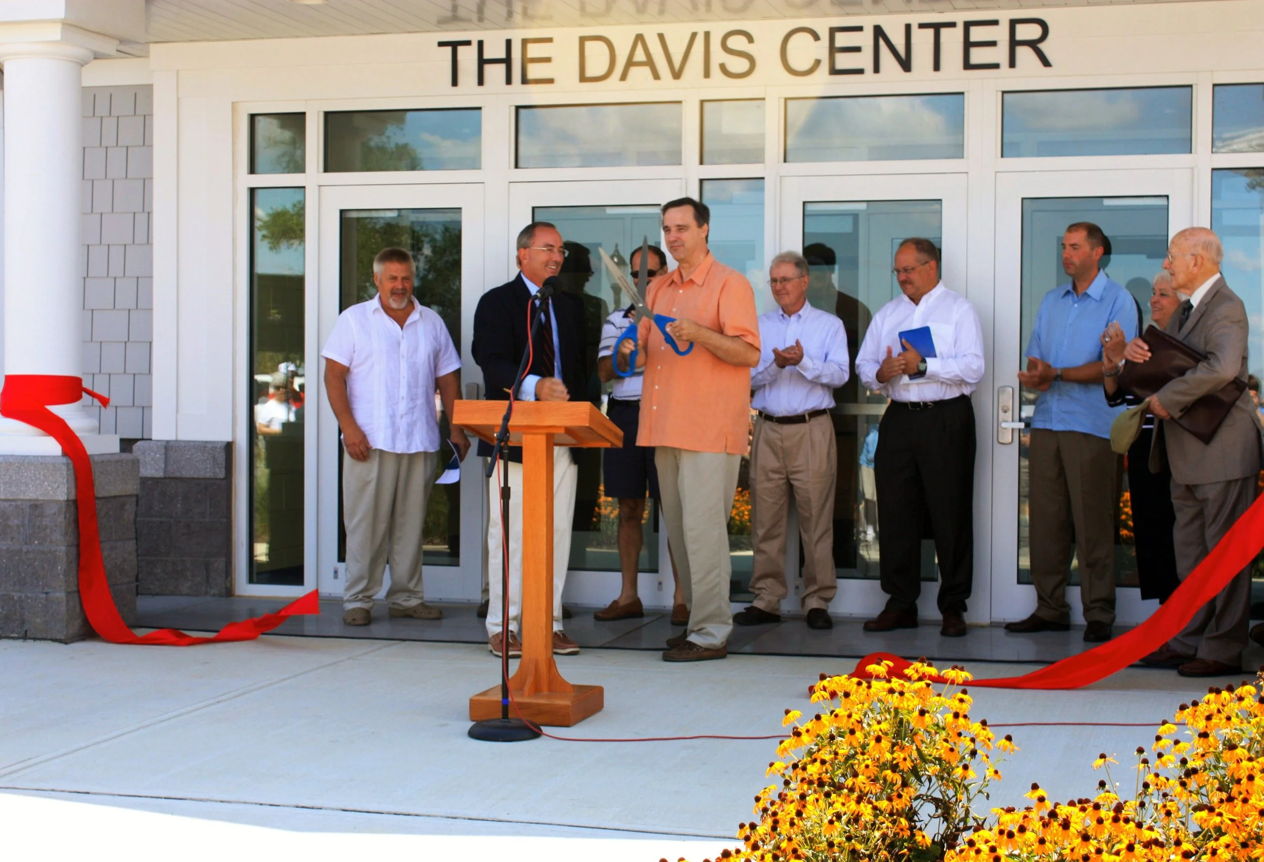 A ribbon-cutting ceremony outside a building named 'The Davis Center' with a small group of people, some holding documents, standing in front of glass doors. There are yellow flowers in the foreground.