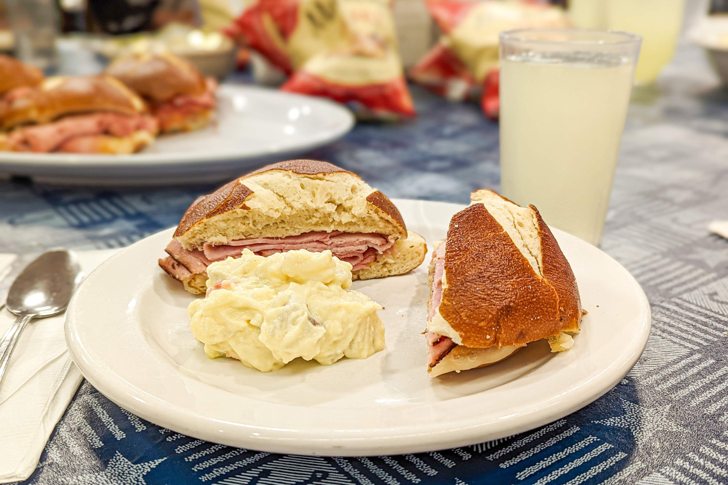 A white plate with a ham sandwich on a hoagie roll, a scoop of potato salad, and a glass of lemonade on a blue tablecloth.