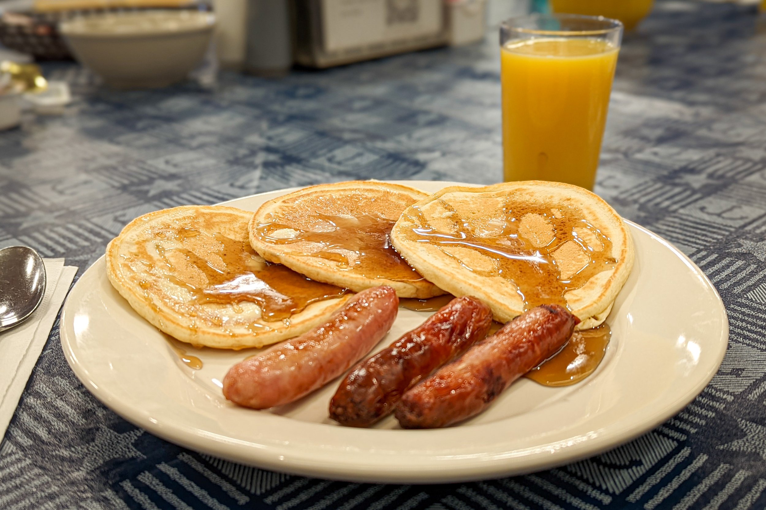Plate of three pancakes with syrup, three breakfast sausages, and a glass of orange juice on a blue tablecloth.