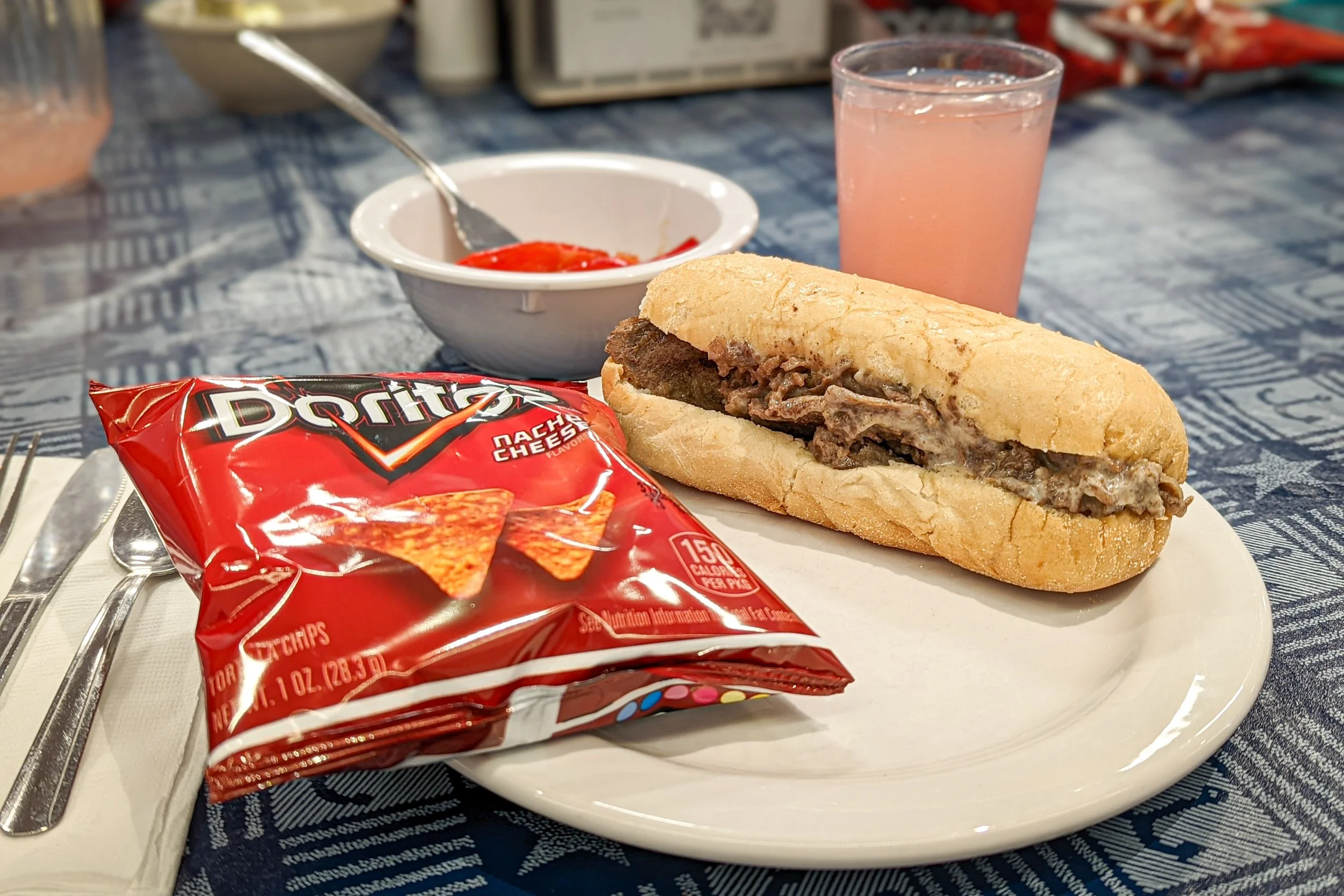 A plate with a beef cheeseburger sub, a bag of Doritos Nacho Cheese chips, a glass of pink lemonade, and a small bowl of ketchup on a blue patterned tablecloth.