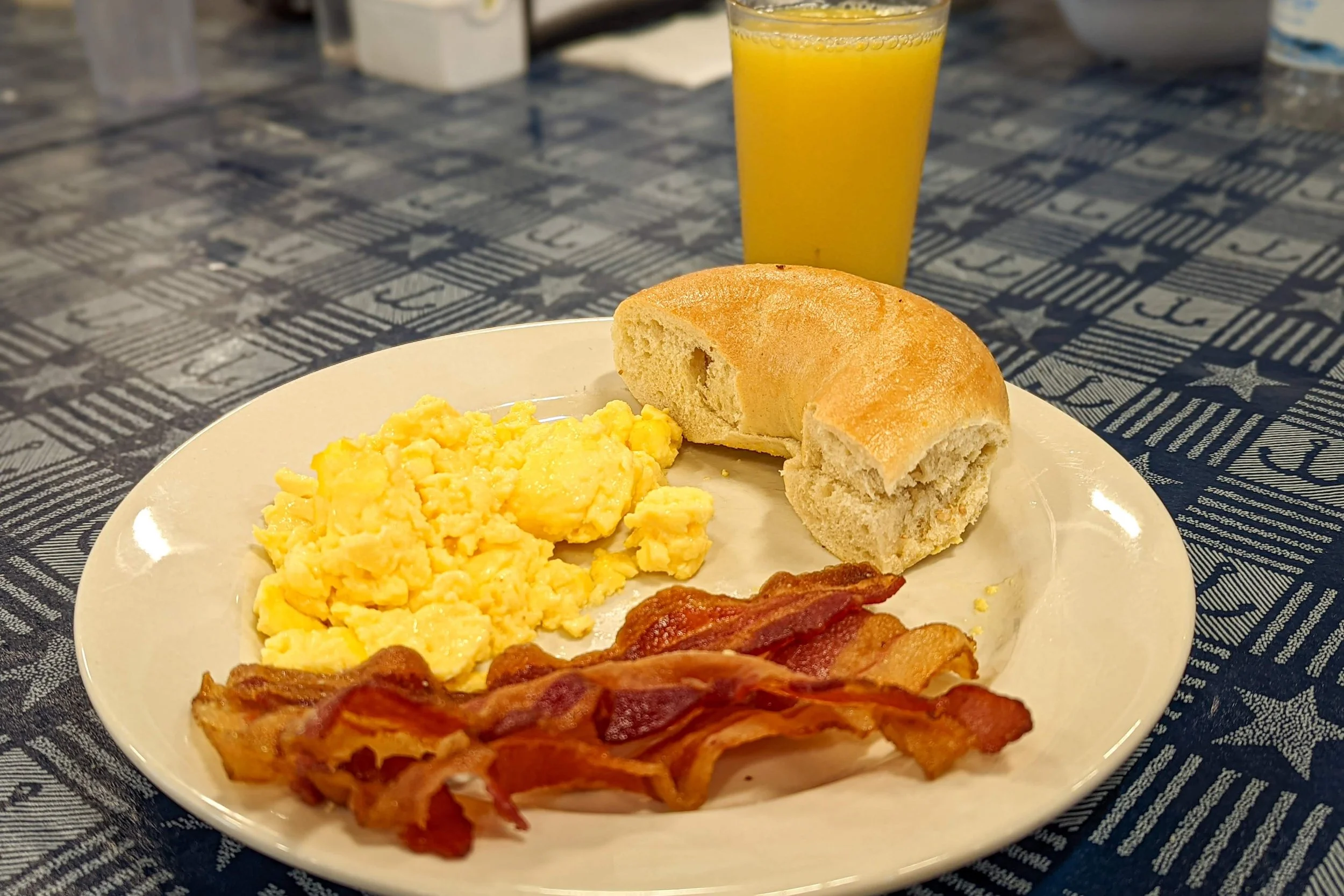 A breakfast plate with scrambled eggs, crispy bacon, a bread roll, and a glass of orange juice on a patriotic-themed tablecloth.