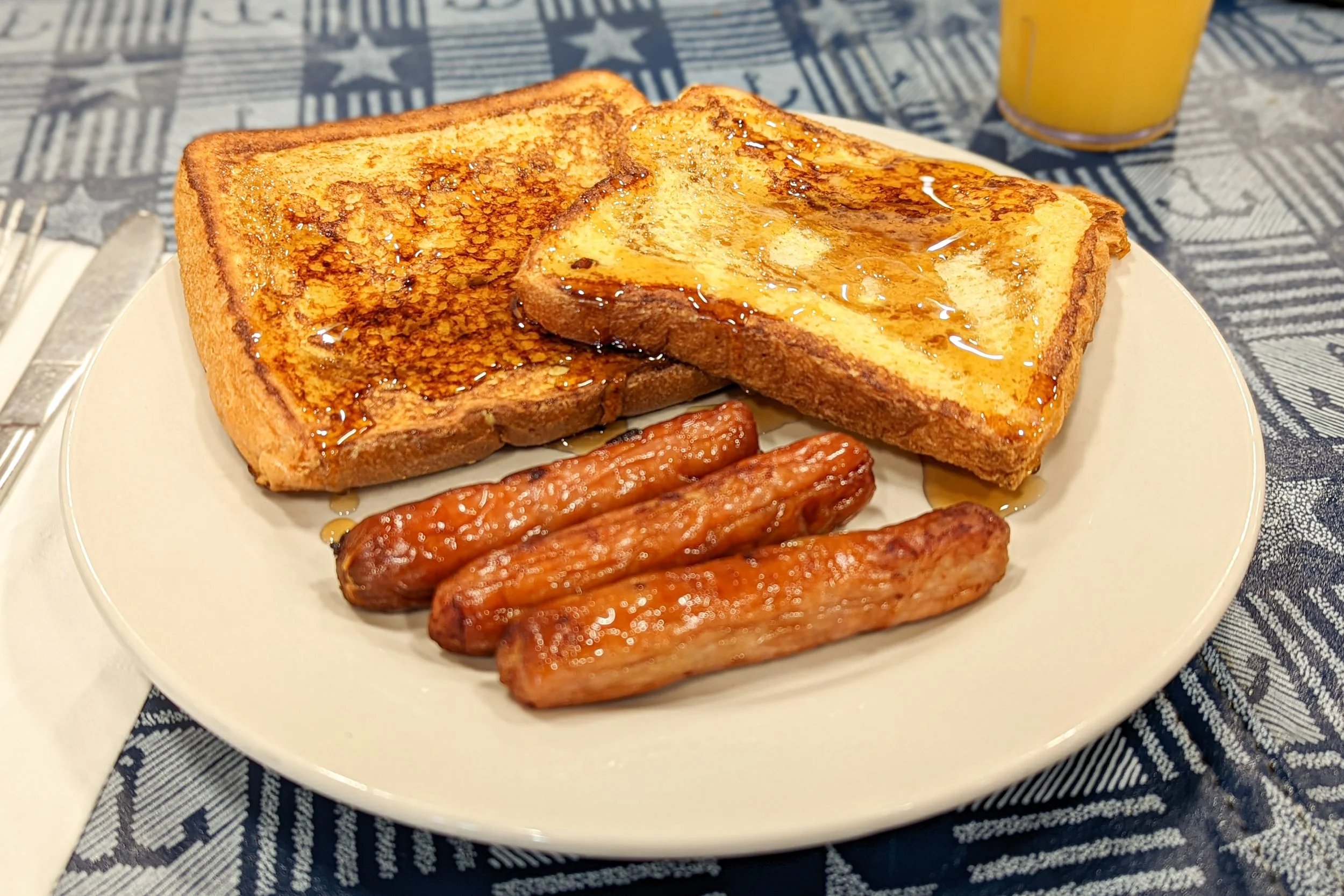 A plate of French toast topped with syrup, accompanied by three sausage links, on a table with a blue patterned tablecloth and a glass of orange juice in the background.