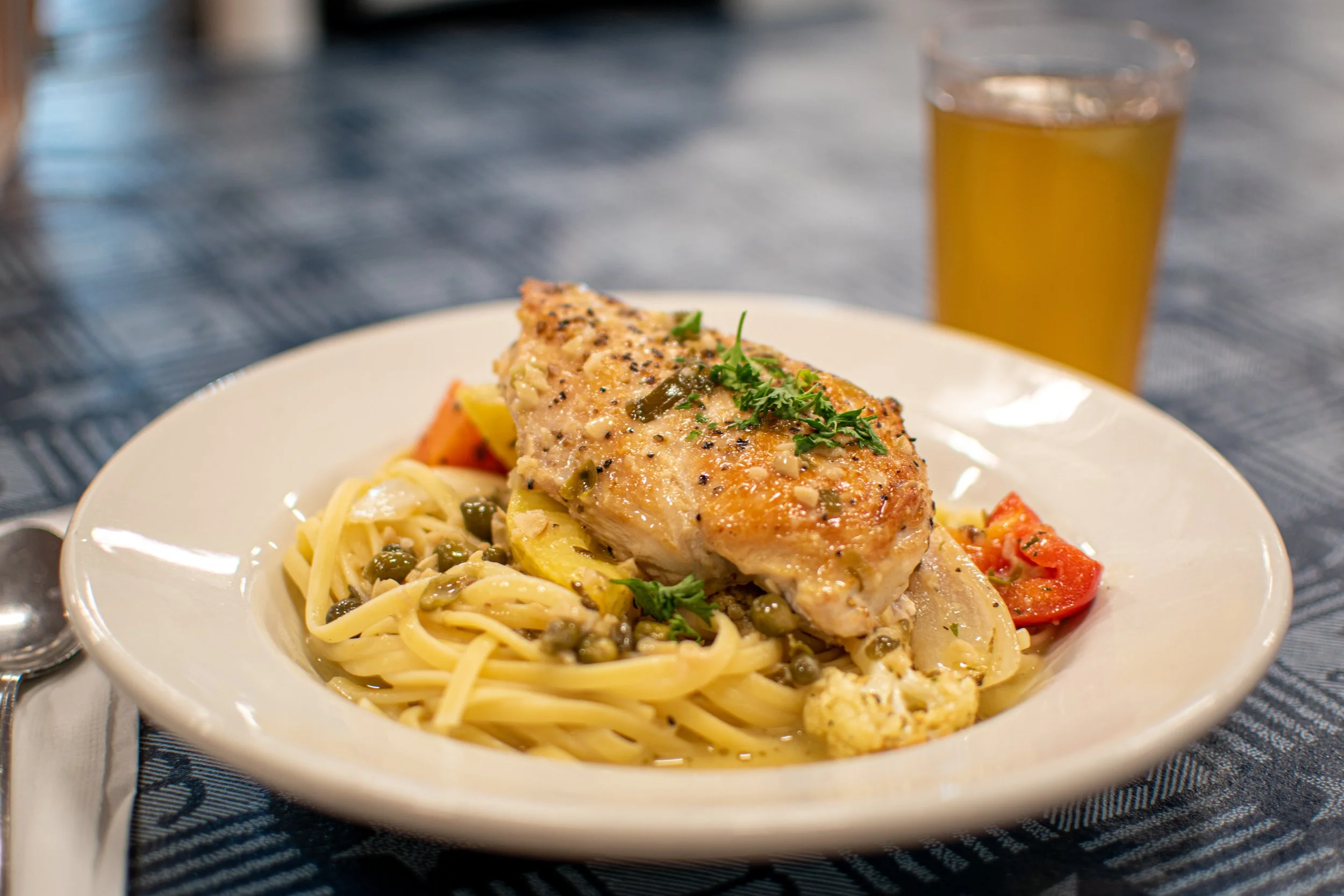 Plate of pasta with cherry tomatoes, capers, and a grilled chicken breast garnished with herbs, with a glass of beer in the background.
