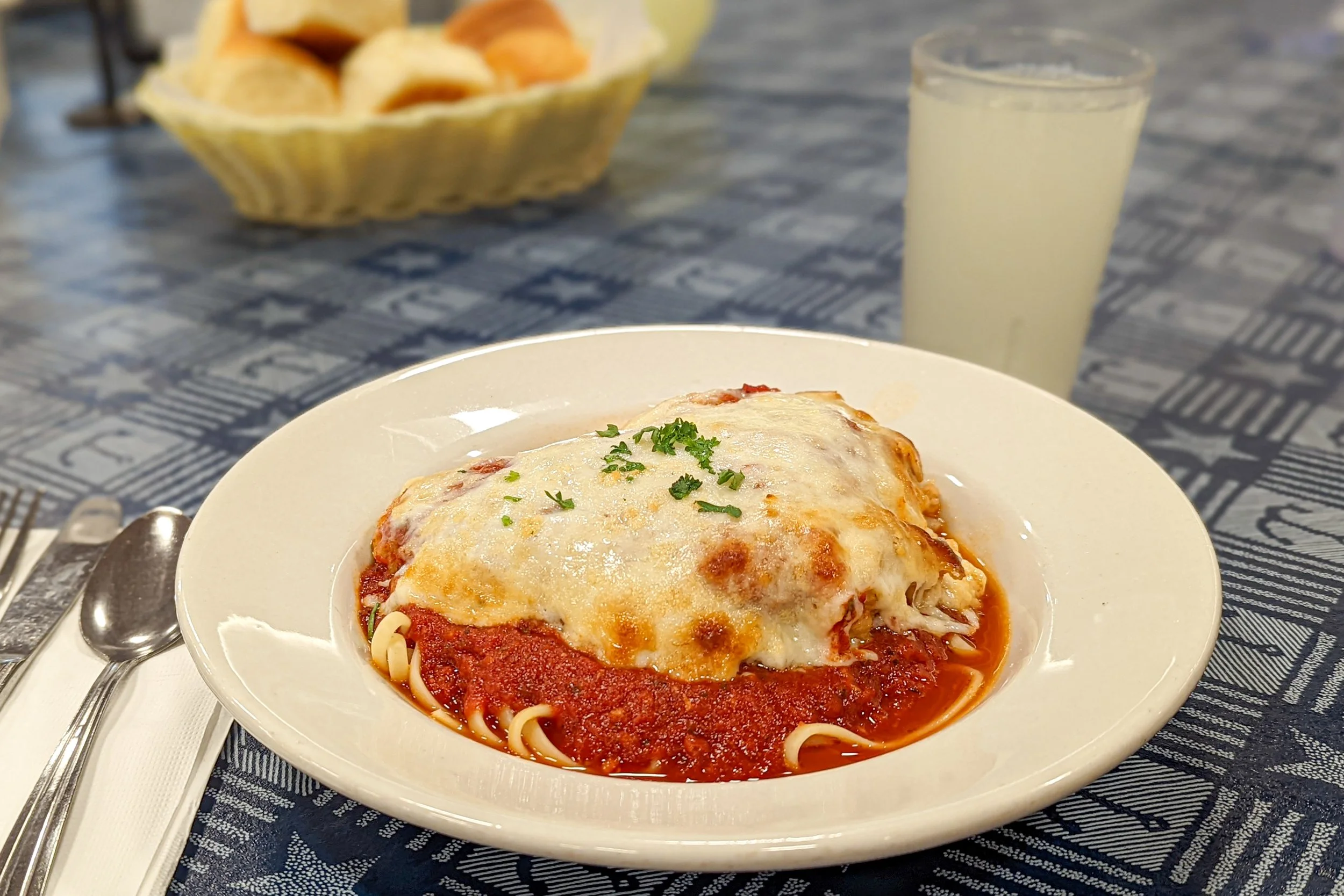 Plate of baked ziti with melted cheese, marinara sauce, and garnished with chopped herbs, on a white plate. In the background, a glass of lemonade and a basket of bread rolls on a blue patterned tablecloth.