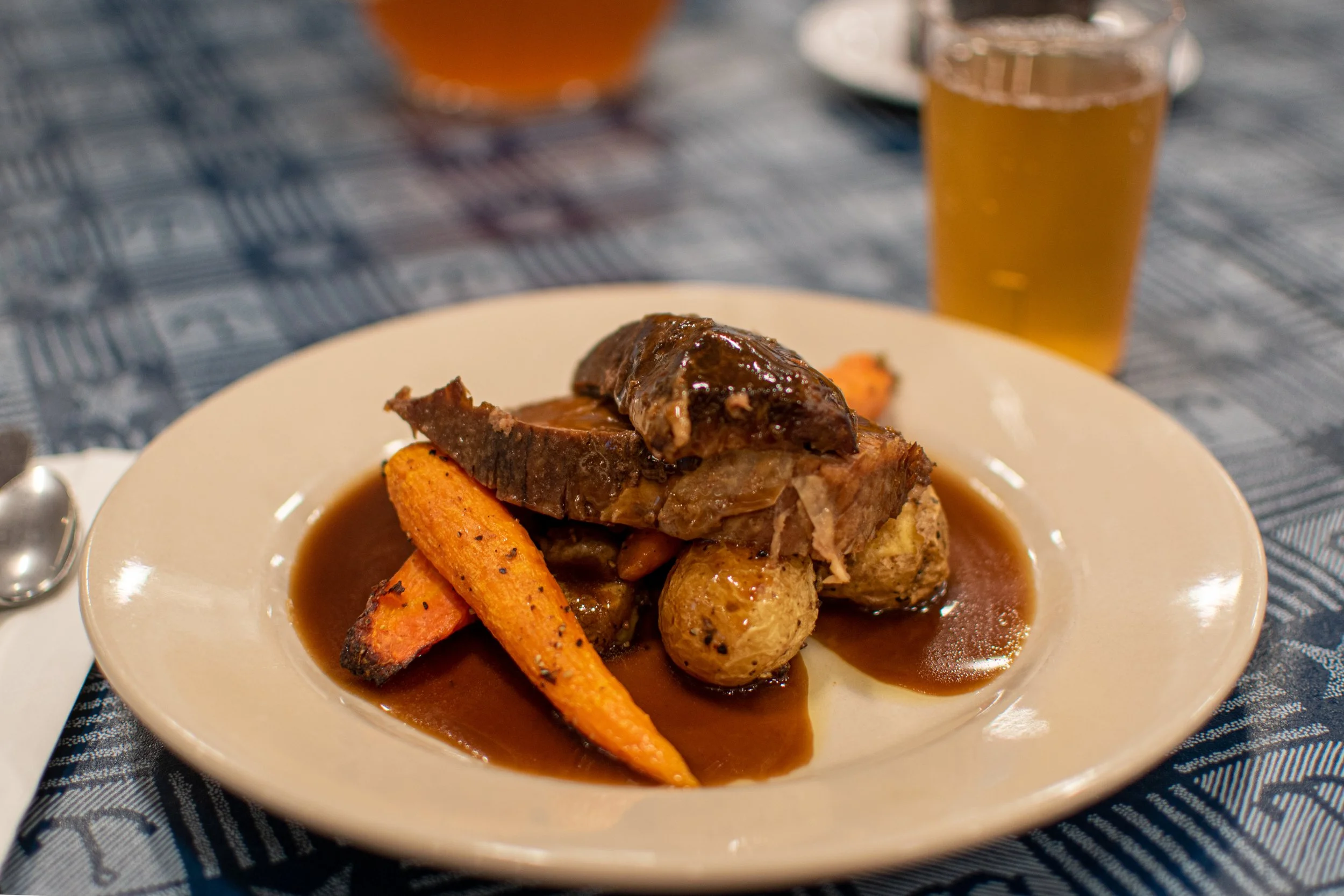 Plate of cooked meat with vegetables and brown gravy, with a glass of beer in the background, on a patterned tablecloth.