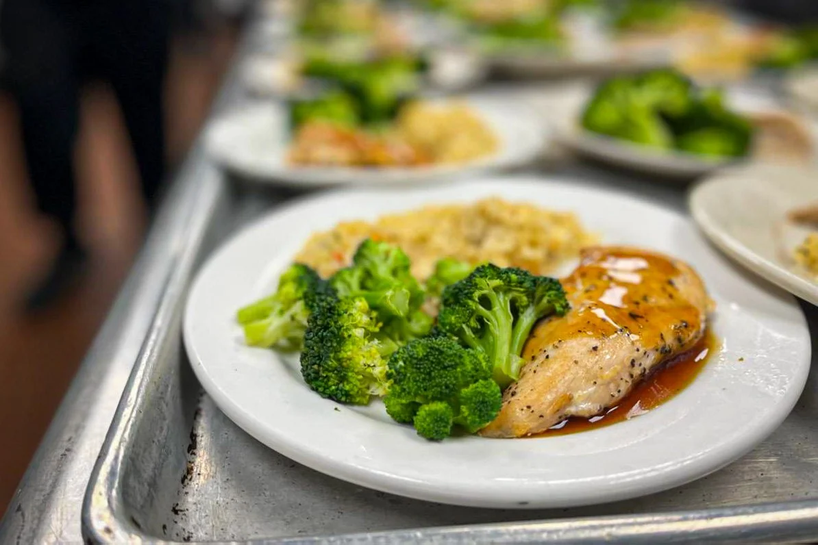 A white plate with cooked broccoli florets, a piece of chicken breast with orange sauce, and a side of rice or grains, on a metal tray.