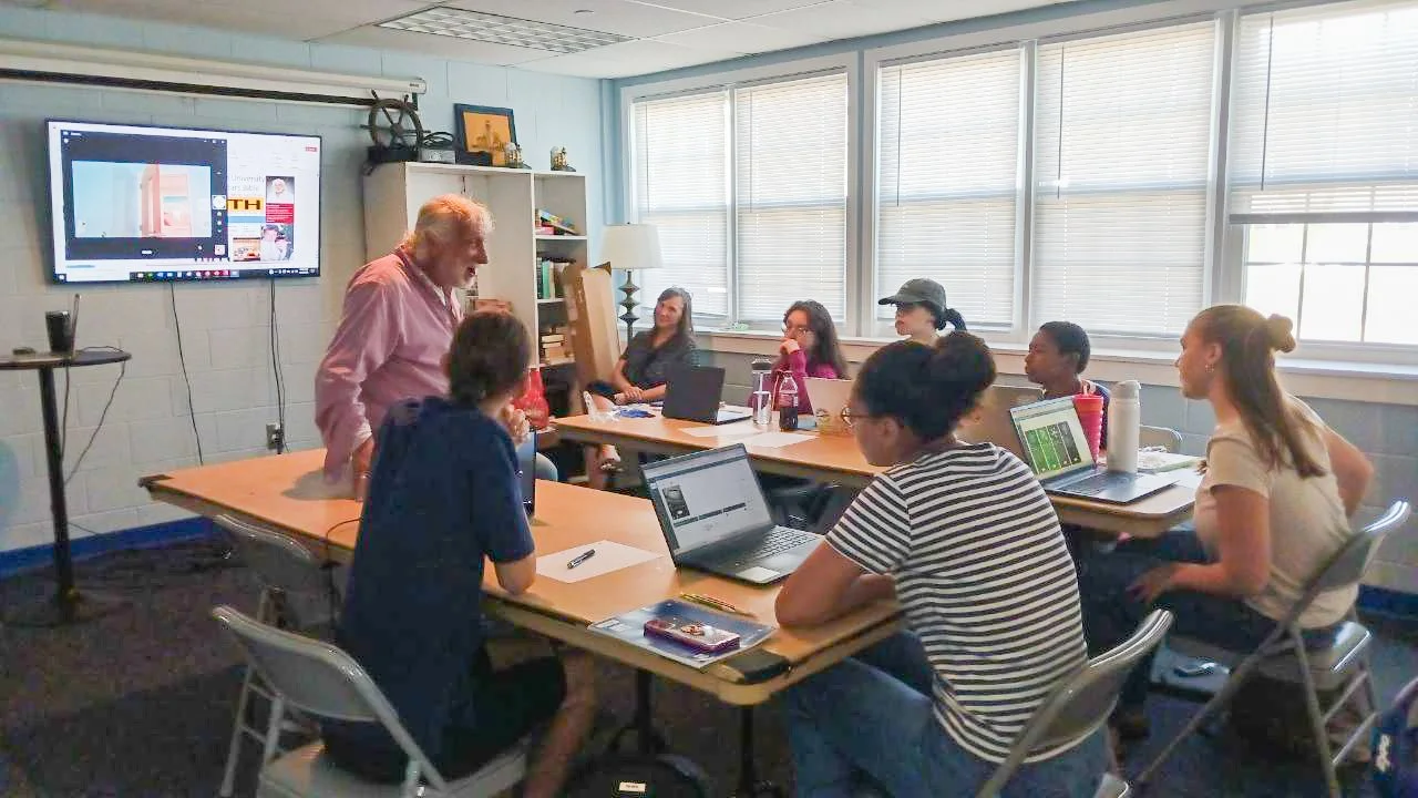 A diverse group of students and an instructor are in a classroom, engaged in a discussion. The students have laptops and notebooks on the table, and a large screen displays a presentation at the front of the room.