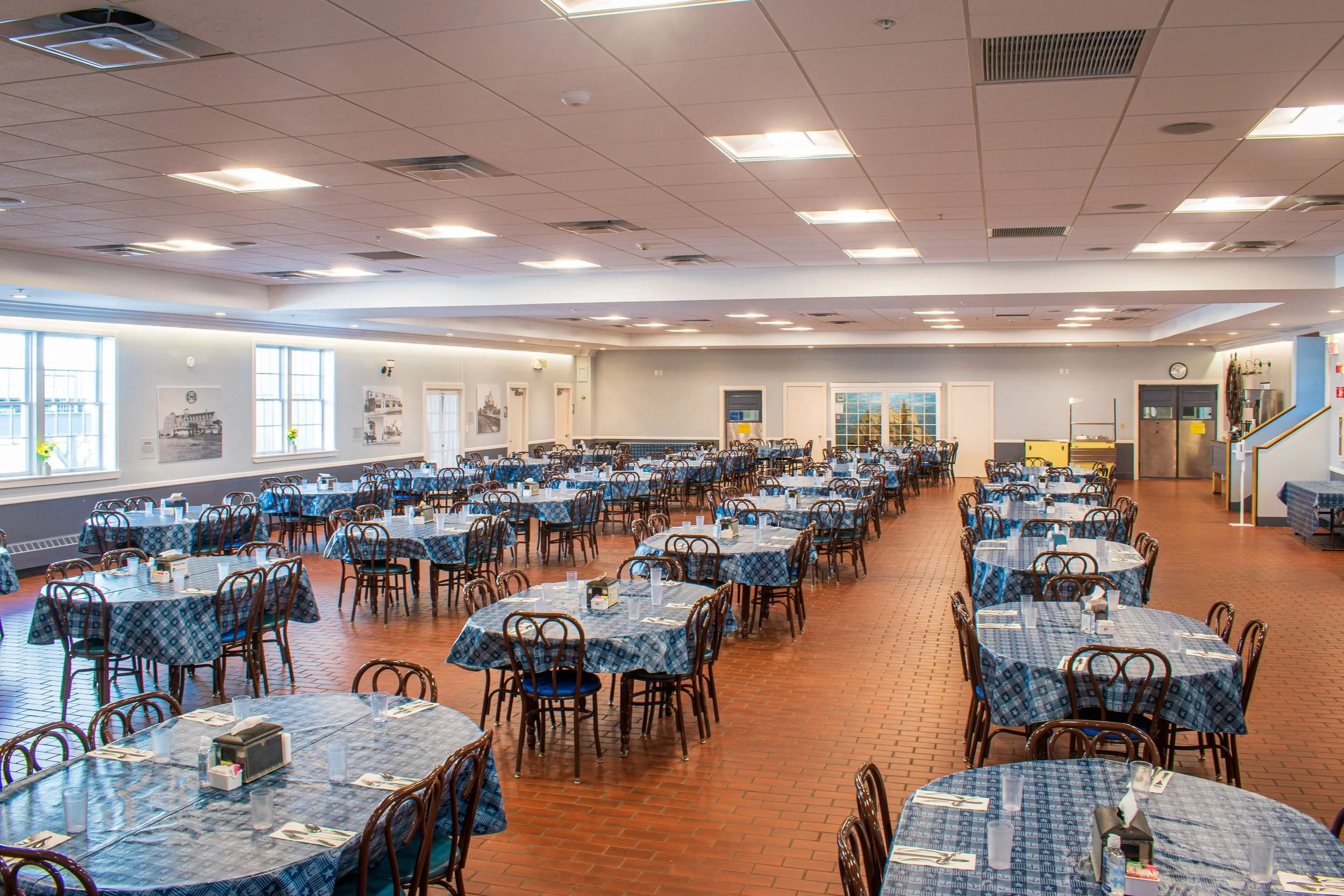 A spacious dining hall with numerous round tables covered with blue and white checkered tablecloths, set with glasses, napkins, and condiments. The room has large windows on one side, with framed black and white photographs hanging on the walls, and wooden chairs surrounding each table.