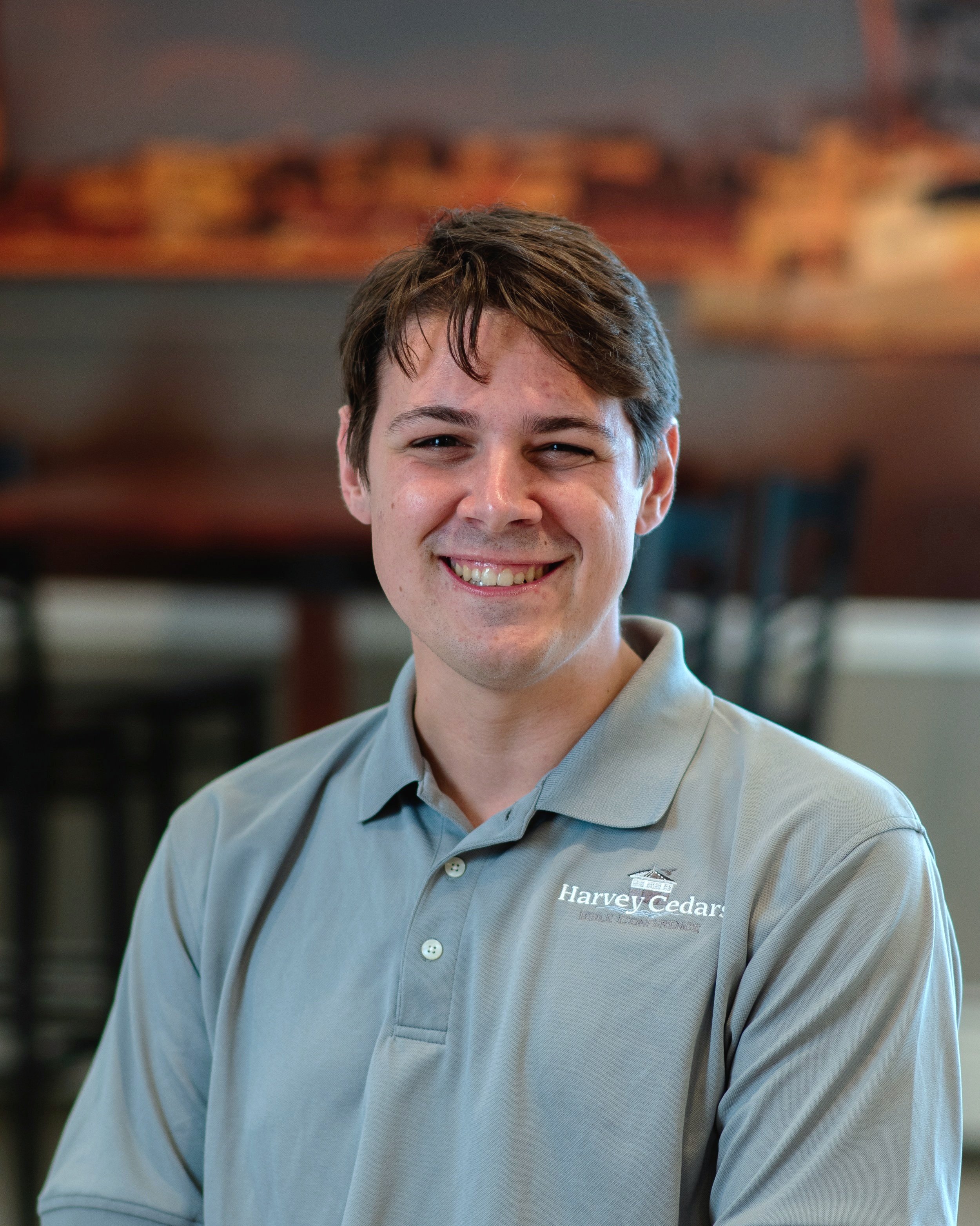 A smiling young man with brown hair wearing a light grey polo shirt with a logo that says 'Harvey Cedars' in front of a blurry background of a room with chairs and a painting.