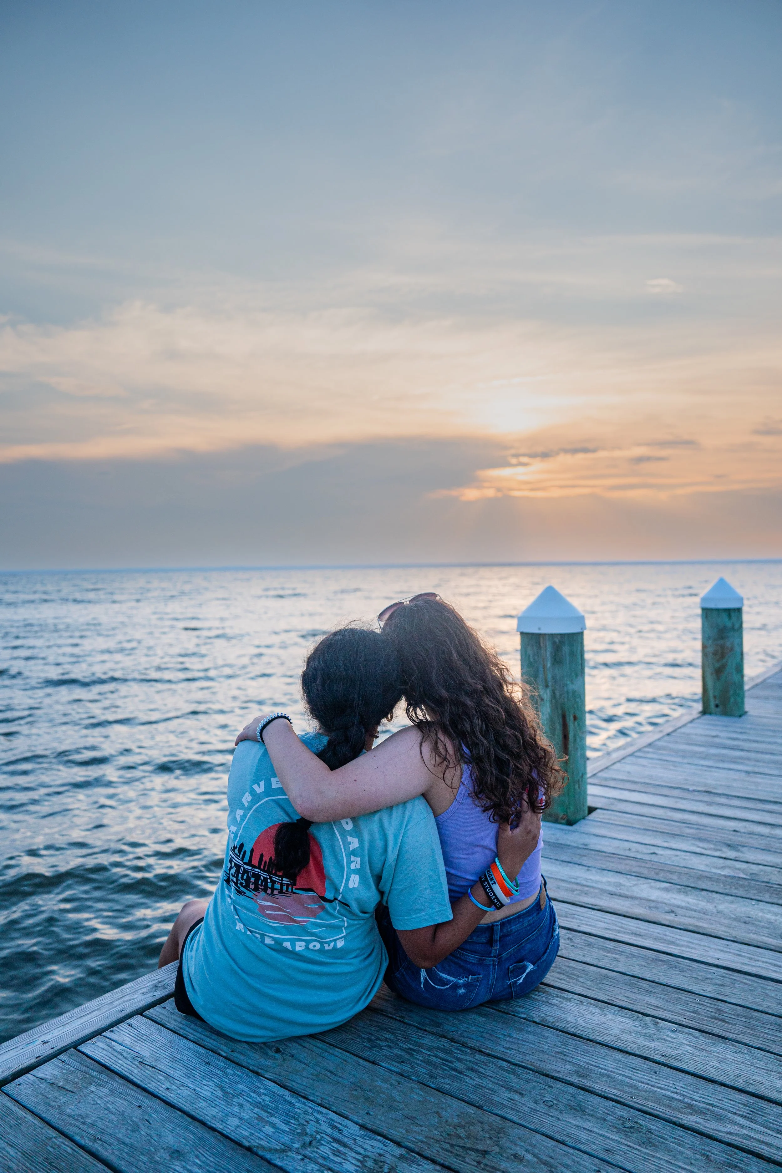 Two girls hugging on a wooden dock by the water at sunset with a sky full of clouds.