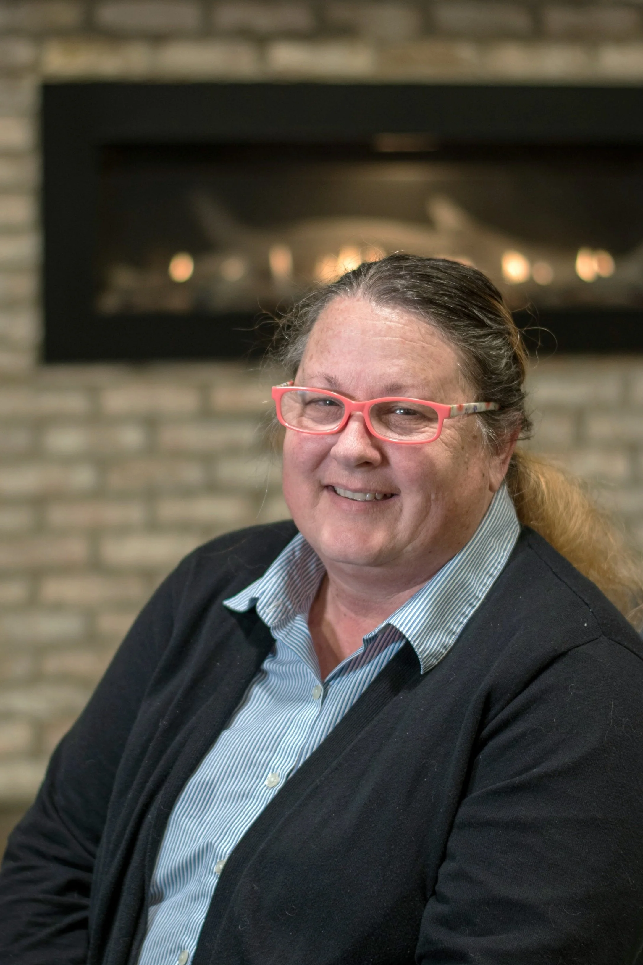 A smiling woman with glasses sitting in front of a brick fireplace.