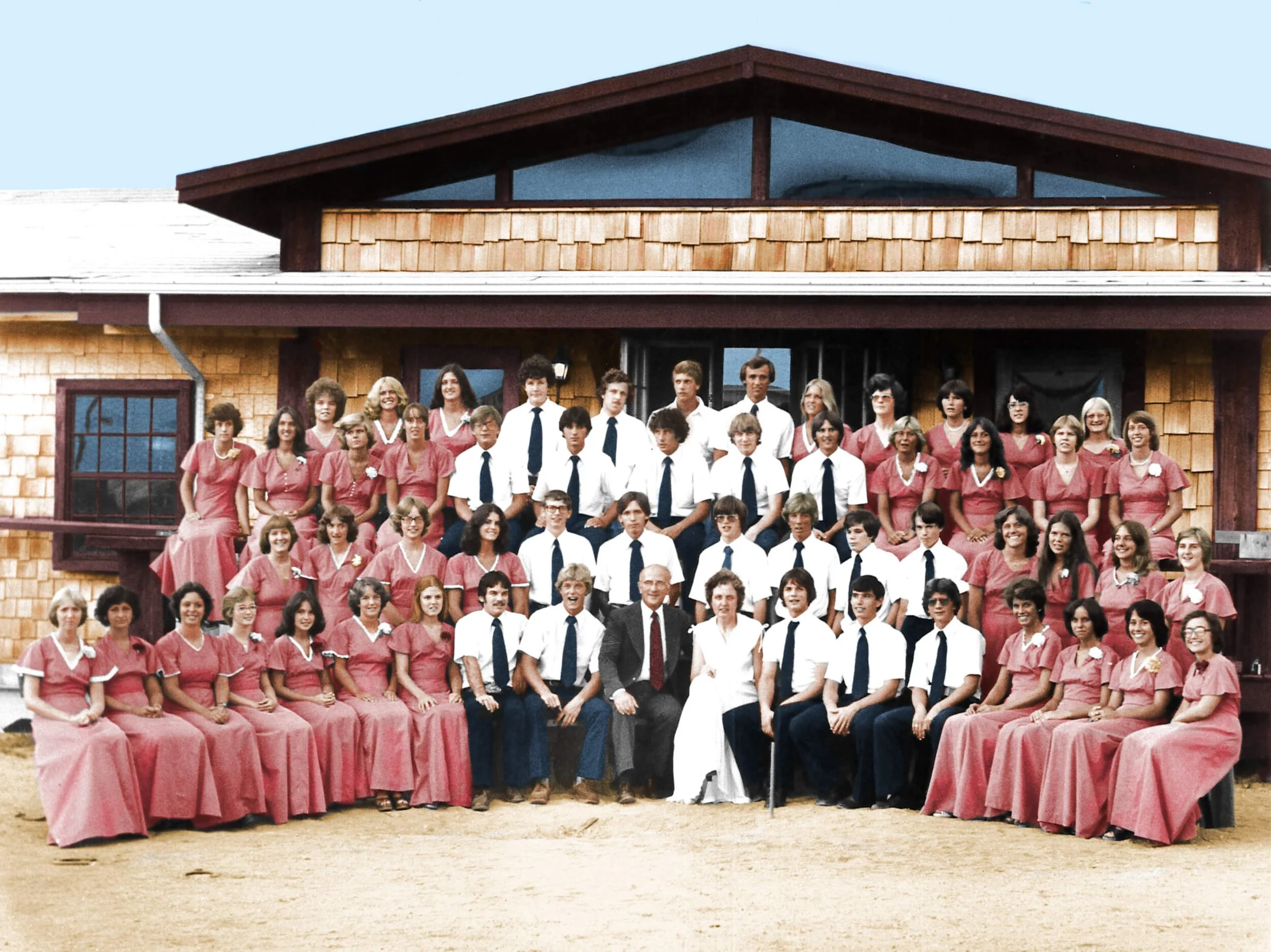 A large group photo of men and women, some in pink dresses and others in white shirts with dark ties, gathered outside a house with a wooden exterior and a sloped roof.