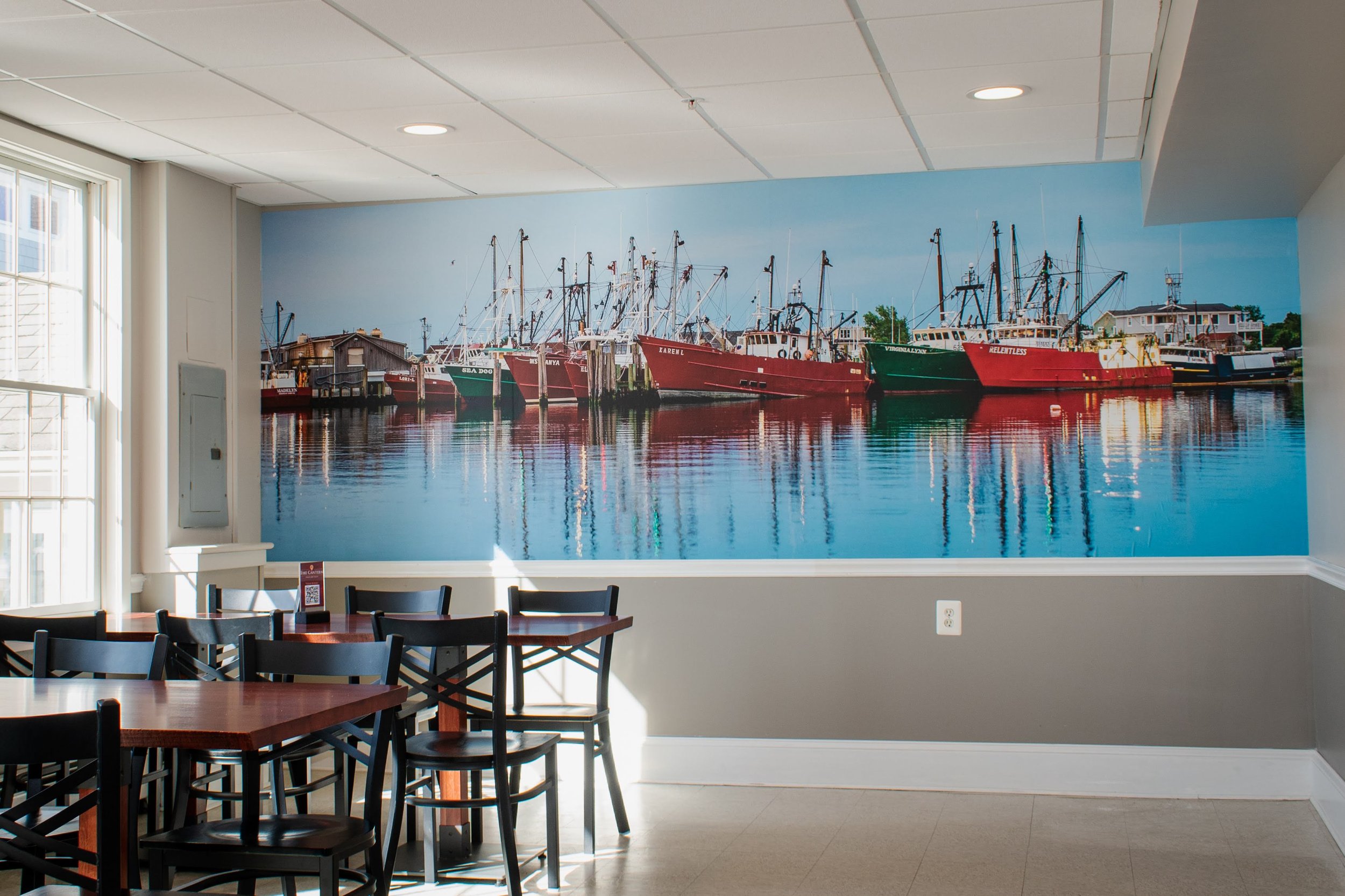 Indoor dining area with tables and chairs, and a large mural of boats docked at a harbor on the wall