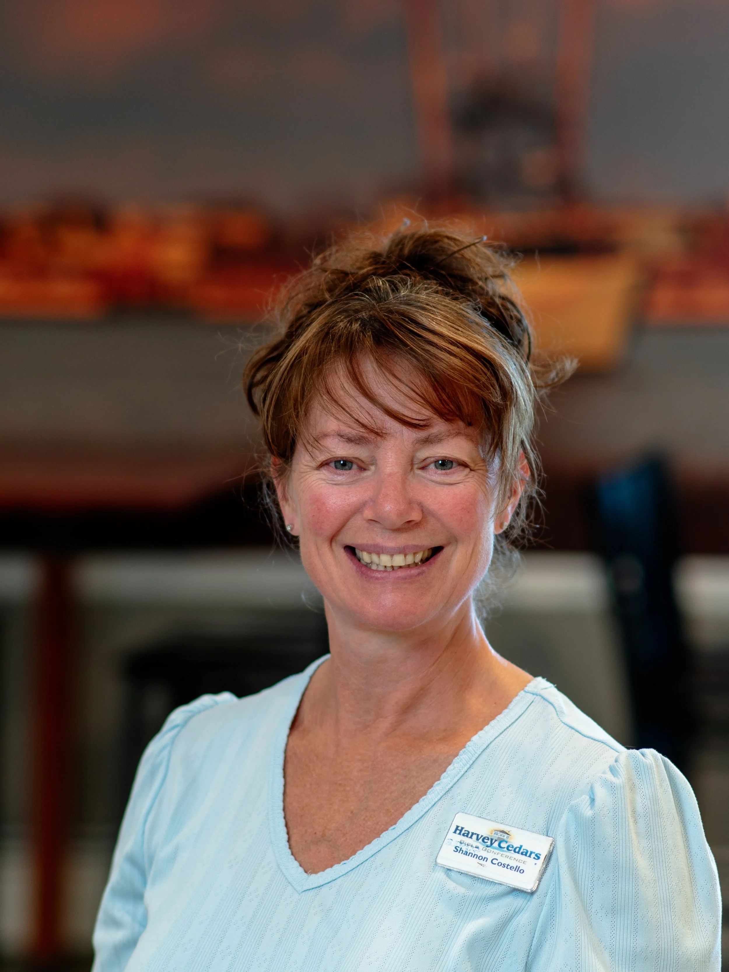 A woman with long brown hair and blue eyes smiling, wearing a blue top, seated indoors with stacked chairs and artwork in the background.