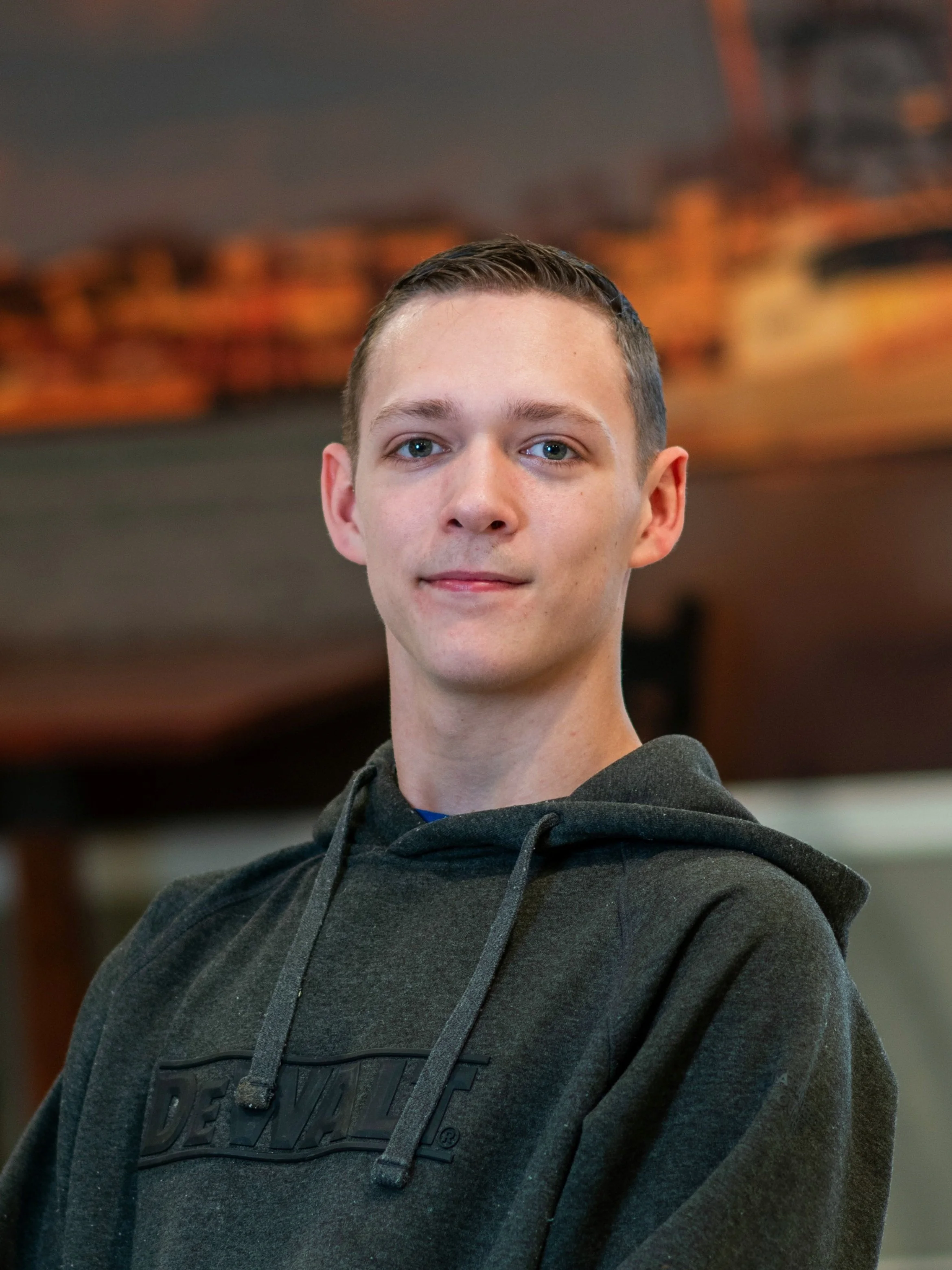 A young man with short brown hair and blue eyes wearing a black hoodie, standing indoors with blurred warm lighting in the background.