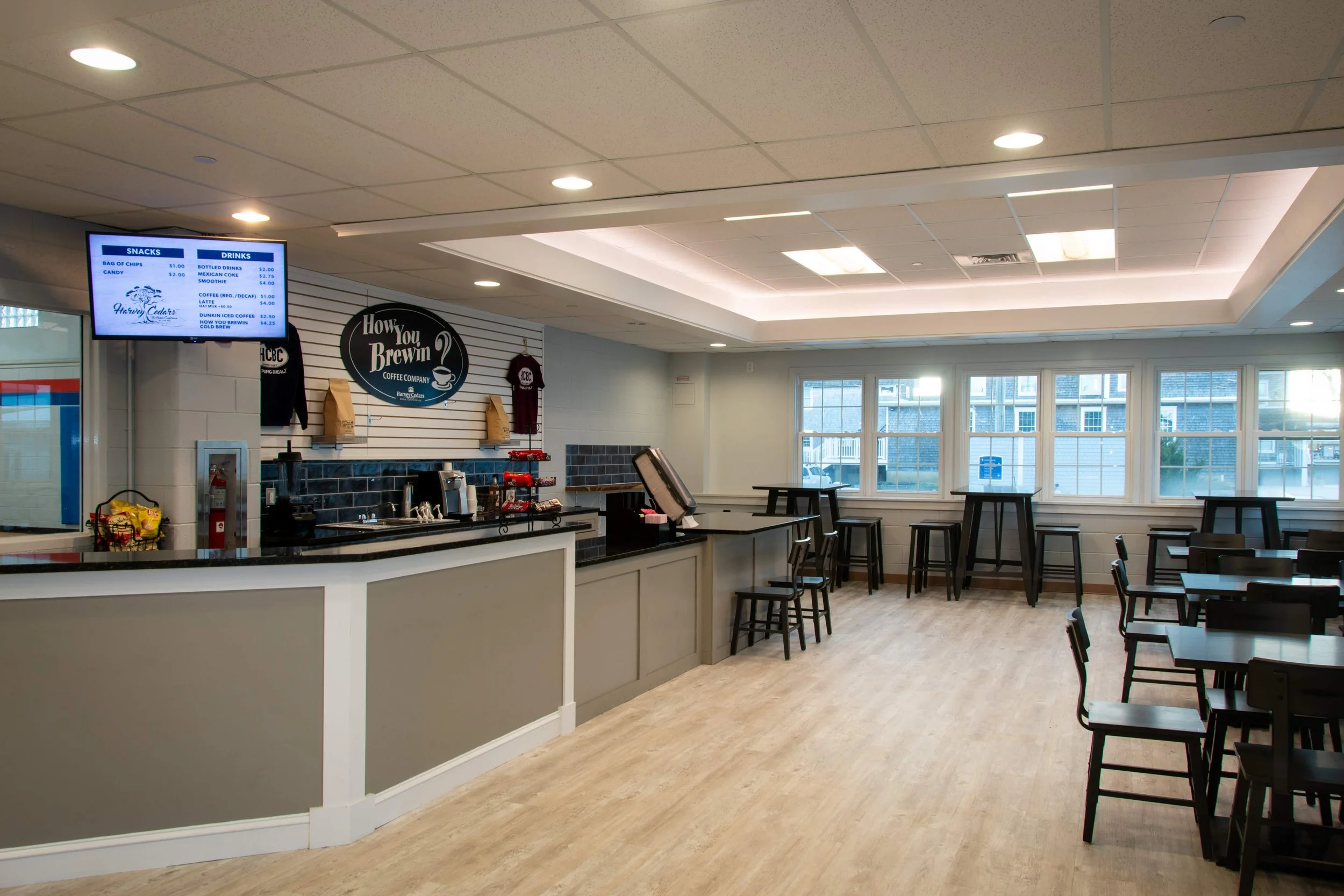 Empty coffee shop with seating area near large windows, a counter with coffee station, digital menu screen on the wall, and minimal decorations.