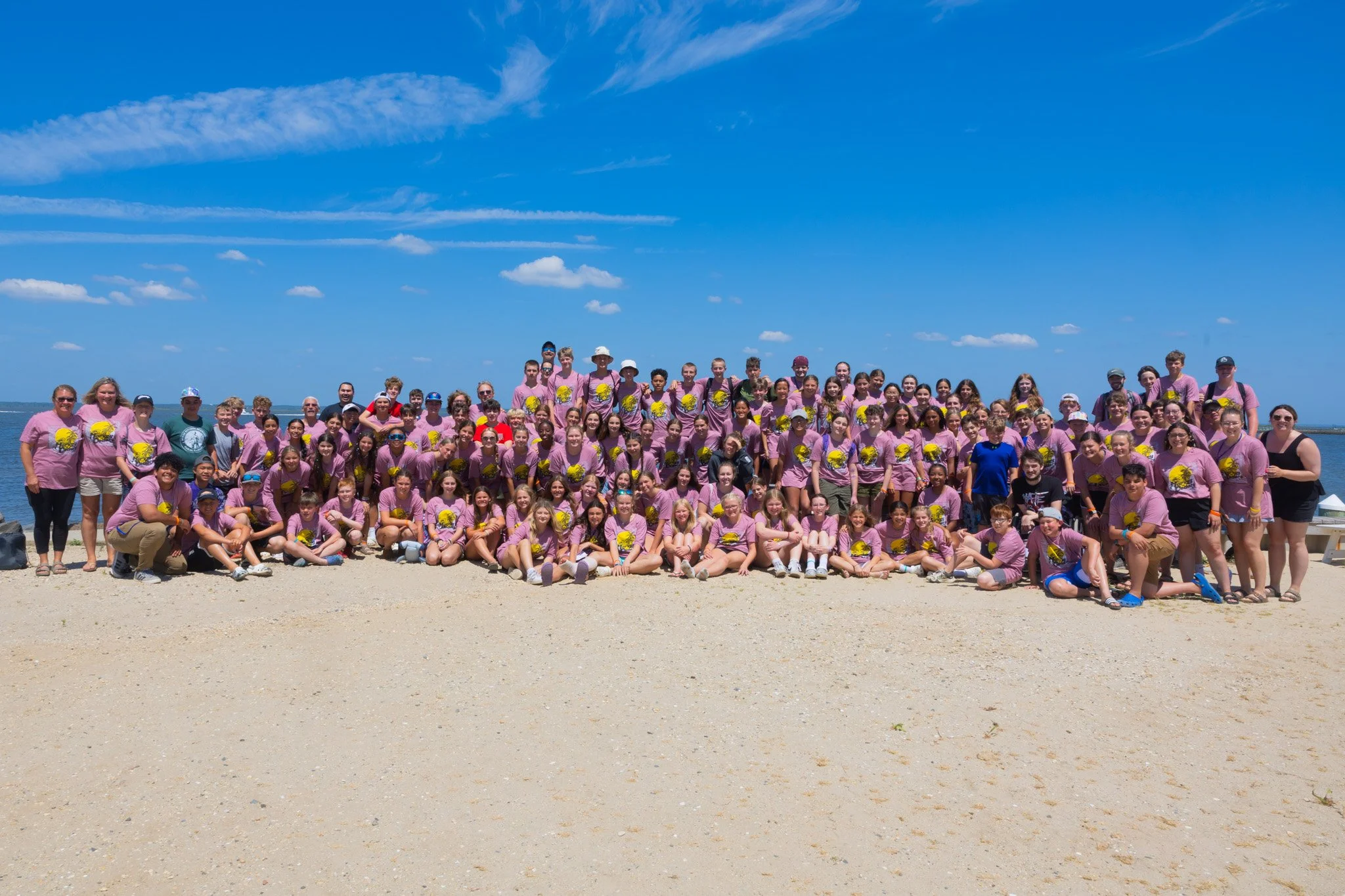 A large group of children and adults at the beach, wearing pink shirts with a yellow graphic on the front, posing for a group photo under a blue sky with a few clouds.