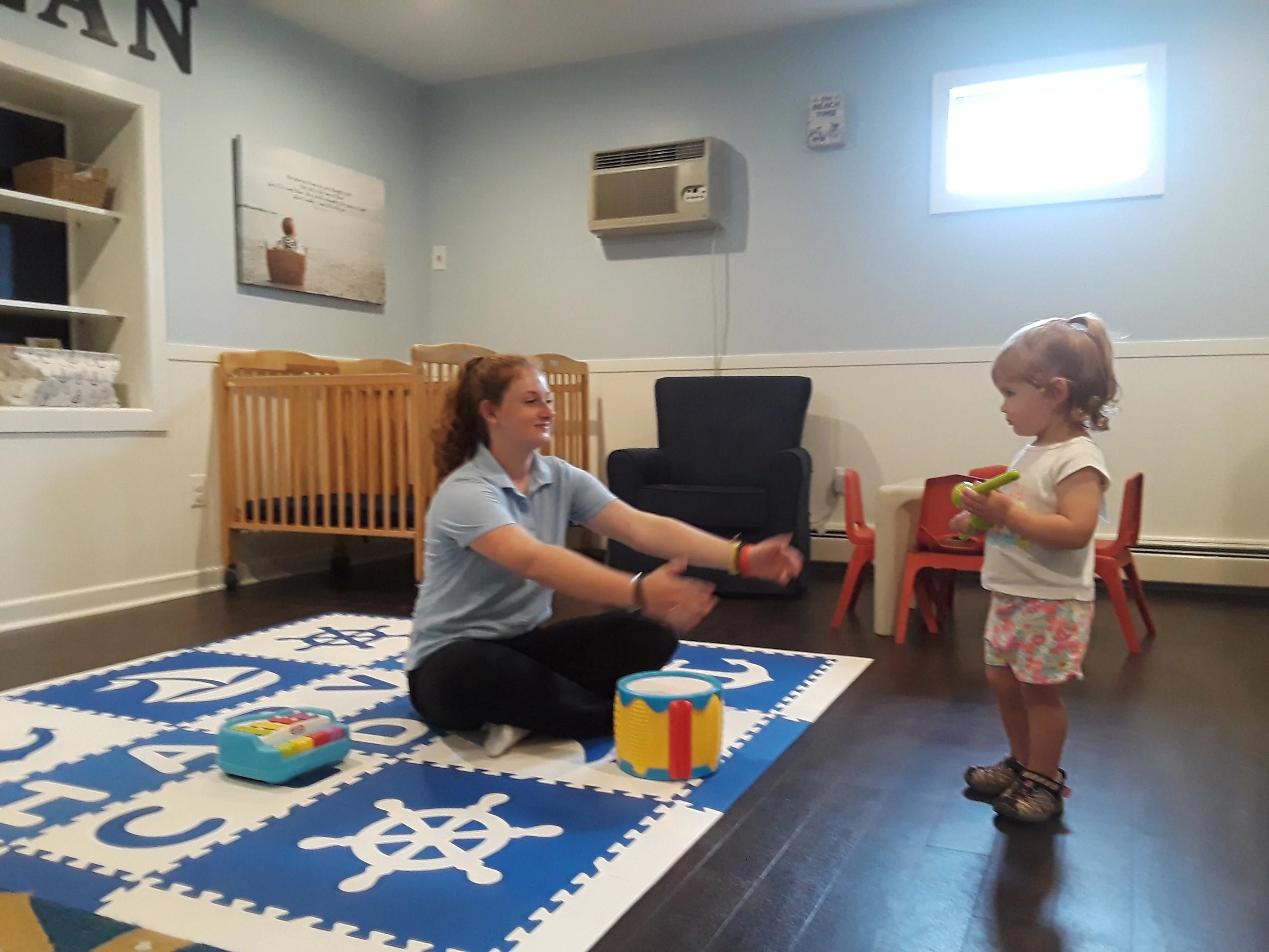 A young woman and a little girl are engaging in a musical activity indoors. The woman is sitting cross-legged on a blue and white nautical-themed foam mat, playing a small drum with a drumstick. The girl stands facing her, holding a green toy tambourine. The room has light blue walls, a small window, and some furniture including a black armchair, a wooden crib, and small red chairs. There is a wall-mounted air conditioner and decorative wall art in the background.