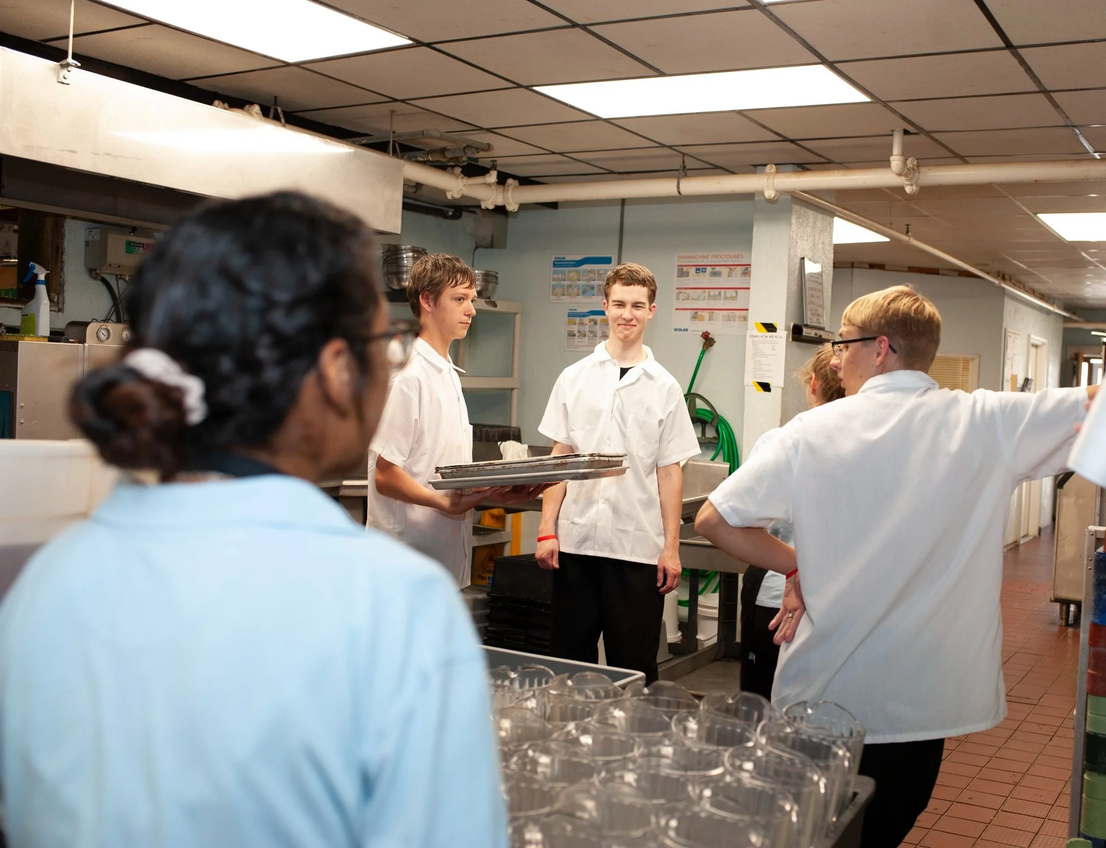 Group of people in kitchen, two young men in white shirts and glasses are talking, one young man is holding trays, a woman with dark hair tied back observe, and others are gathered around in a professional kitchen setting.