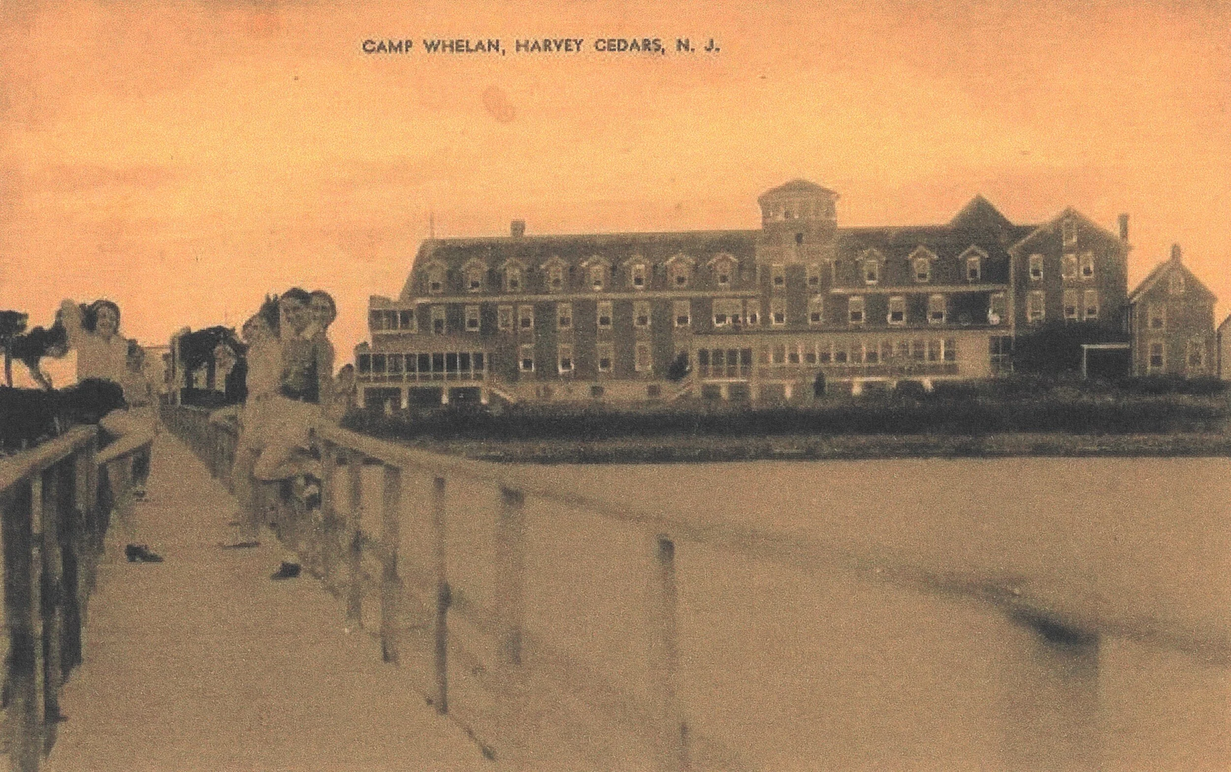 An old sepia-toned photograph of a large, multi-story hotel building called Camp Whelen in Harvey Cedars, New Jersey, viewed from a boardwalk with children leaning on the railing, and trees in the background.