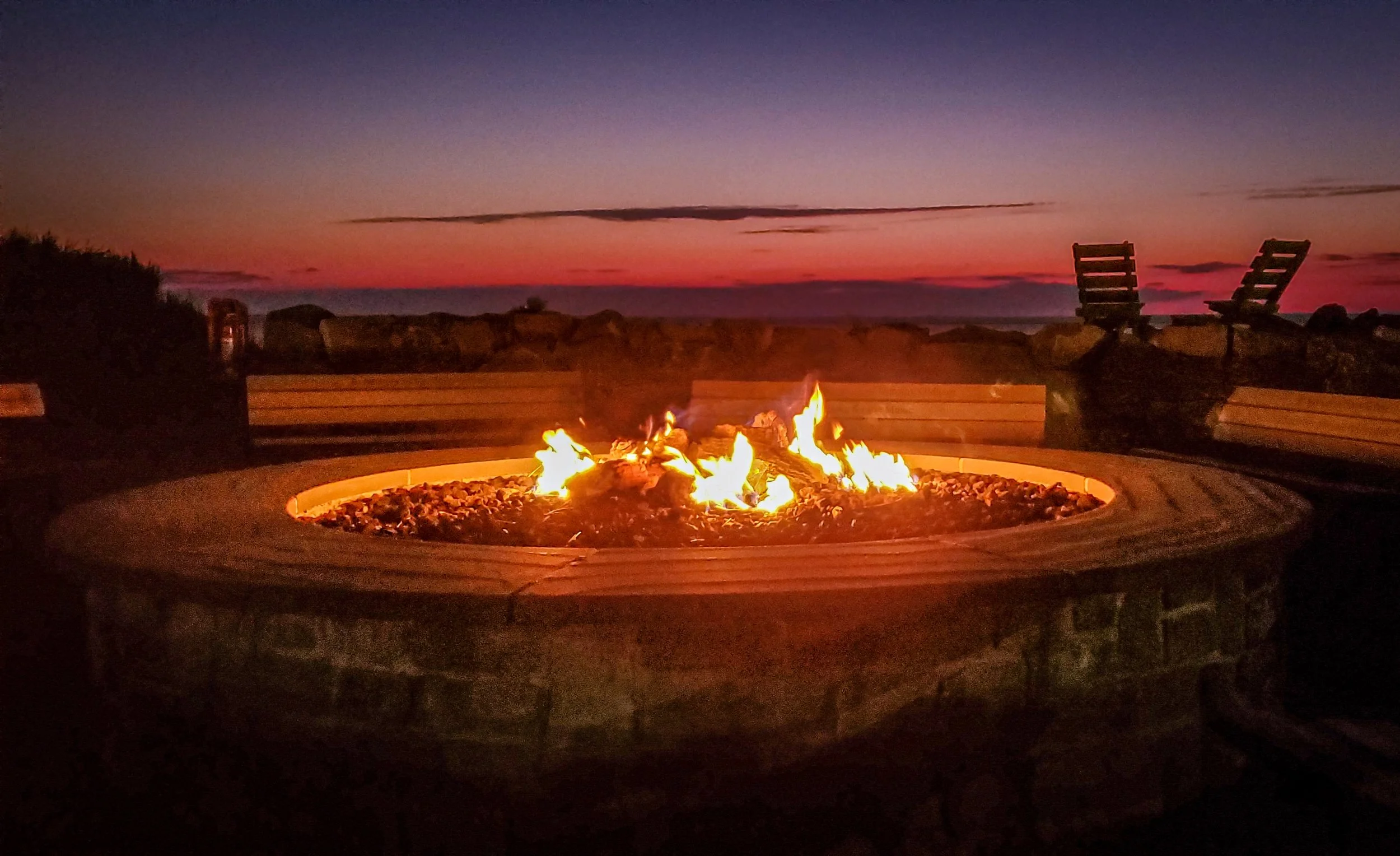 A campfire burning in a circular stone fire pit on a patio at sunset, with a distant view of the sky transitioning from purple to orange and two empty chairs facing the fire.