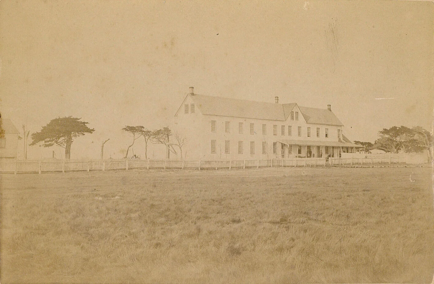 Old sepia photograph of a large, multi-story wooden house with a porch, surrounded by a wooden fence and sparse trees in an open grassy area.