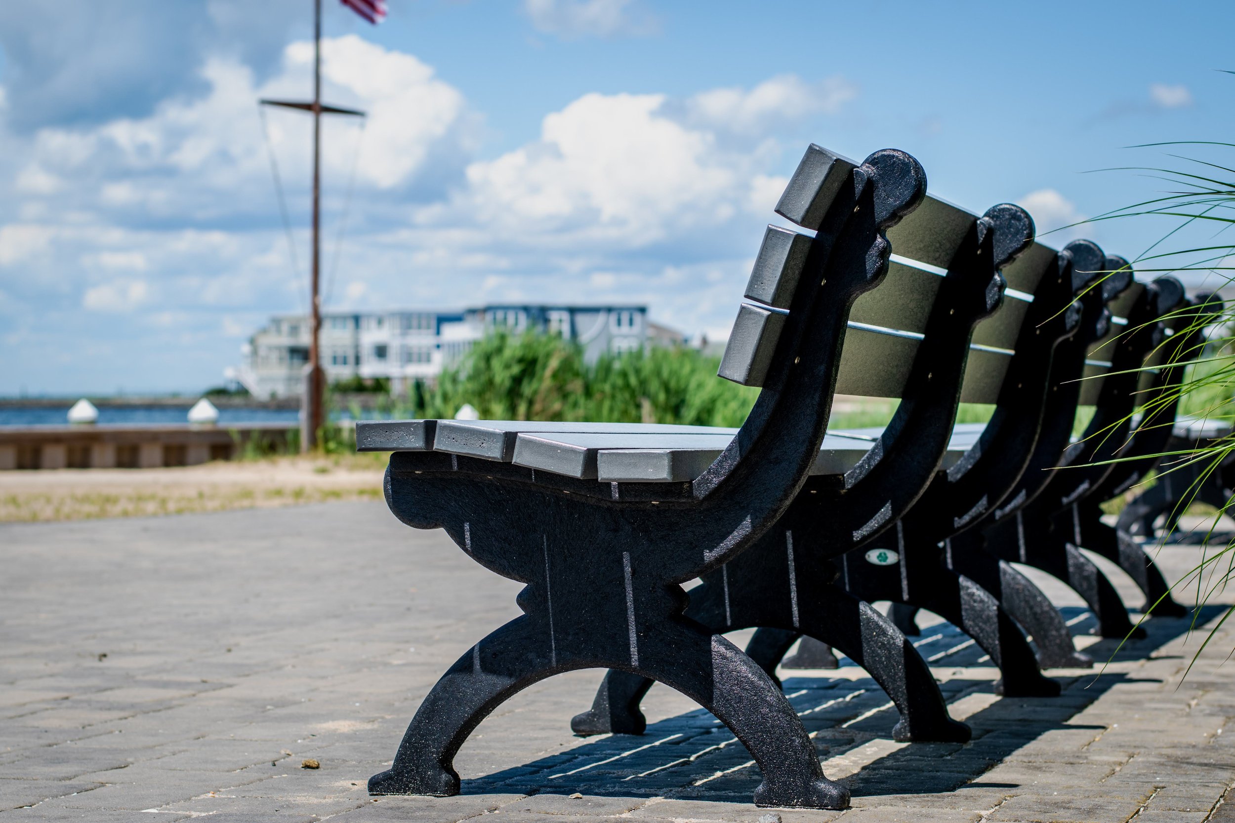A row of empty black park benches on a paved walkway by the water with a cloudy blue sky and a building in the background.
