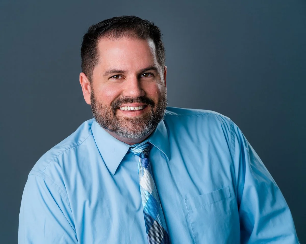 Headshot of a smiling man with dark hair and a beard, wearing a blue dress shirt and a patterned tie, against a gray background.