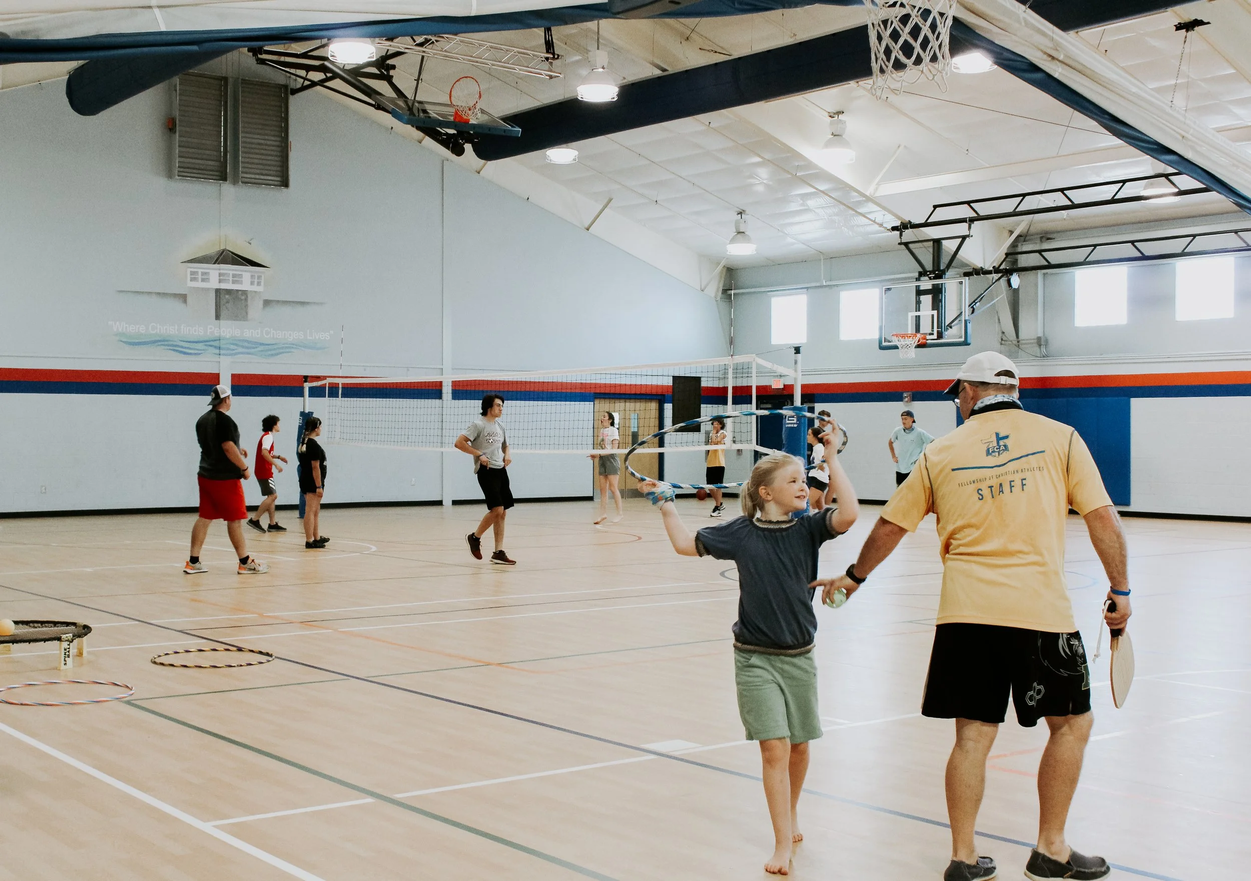 Children and adults playing badminton in an indoor gymnasium with a basketball hoop, volleyball net, and wooden floor.