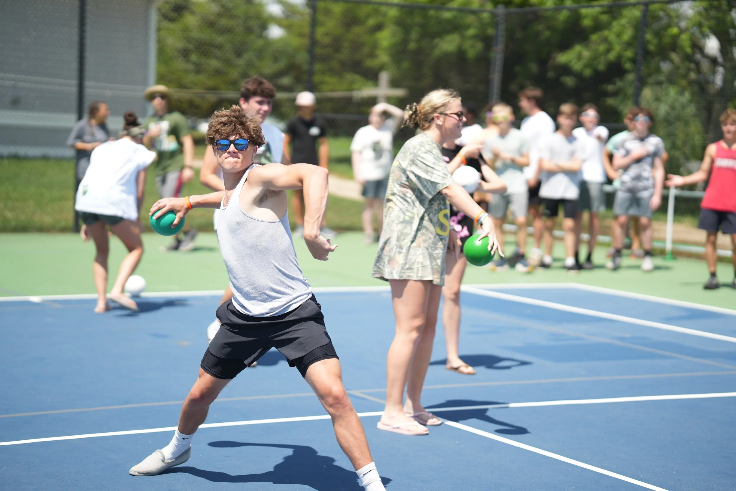 A young man in sunglasses, a white tank top, and black shorts is playing pickleball on an outdoor court, holding a green paddle, with a group of people watching him in the background.