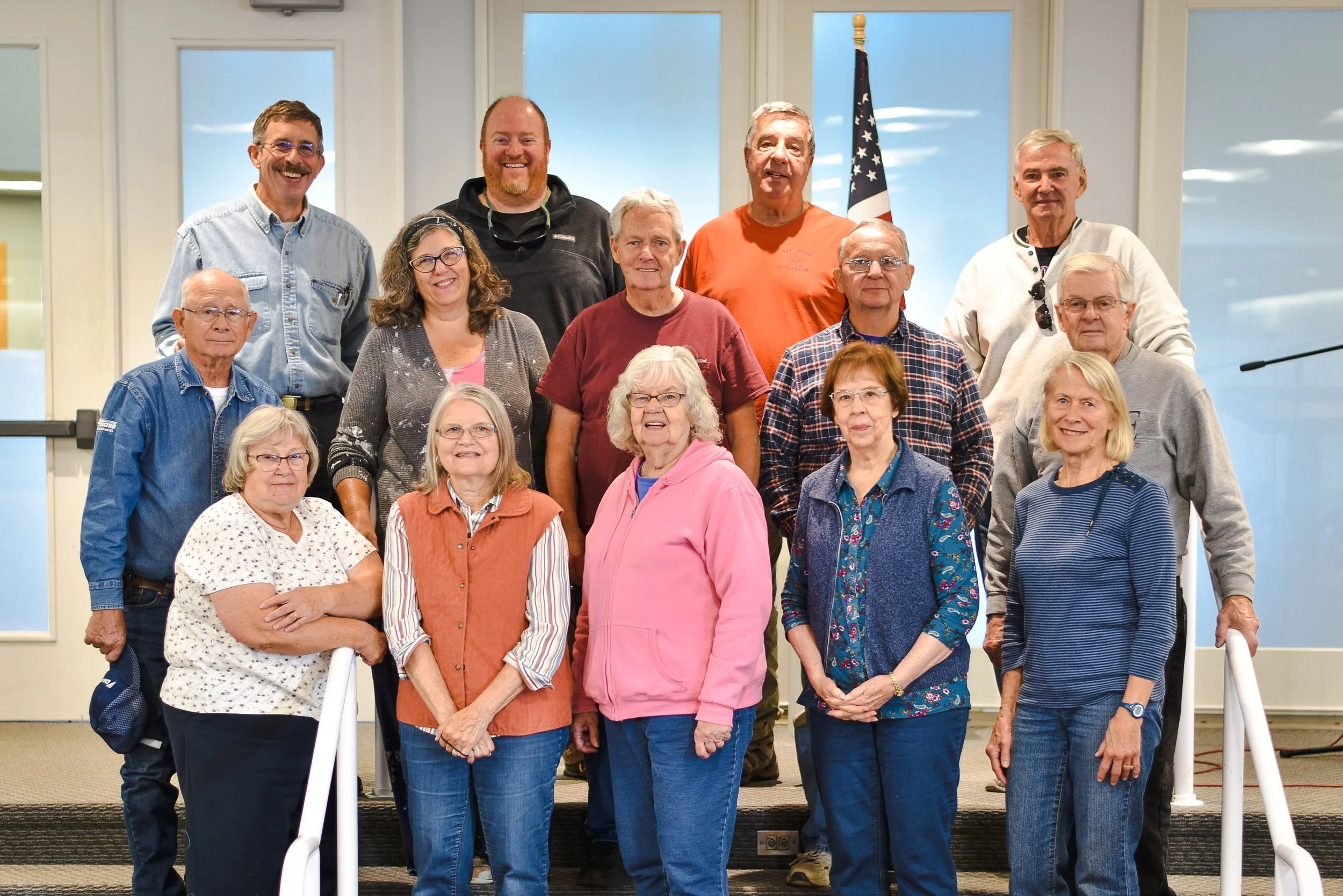 Group photo of fifteen individuals, including men and women of varying ages, standing on a staircase inside a building with windows and an American flag in the background.