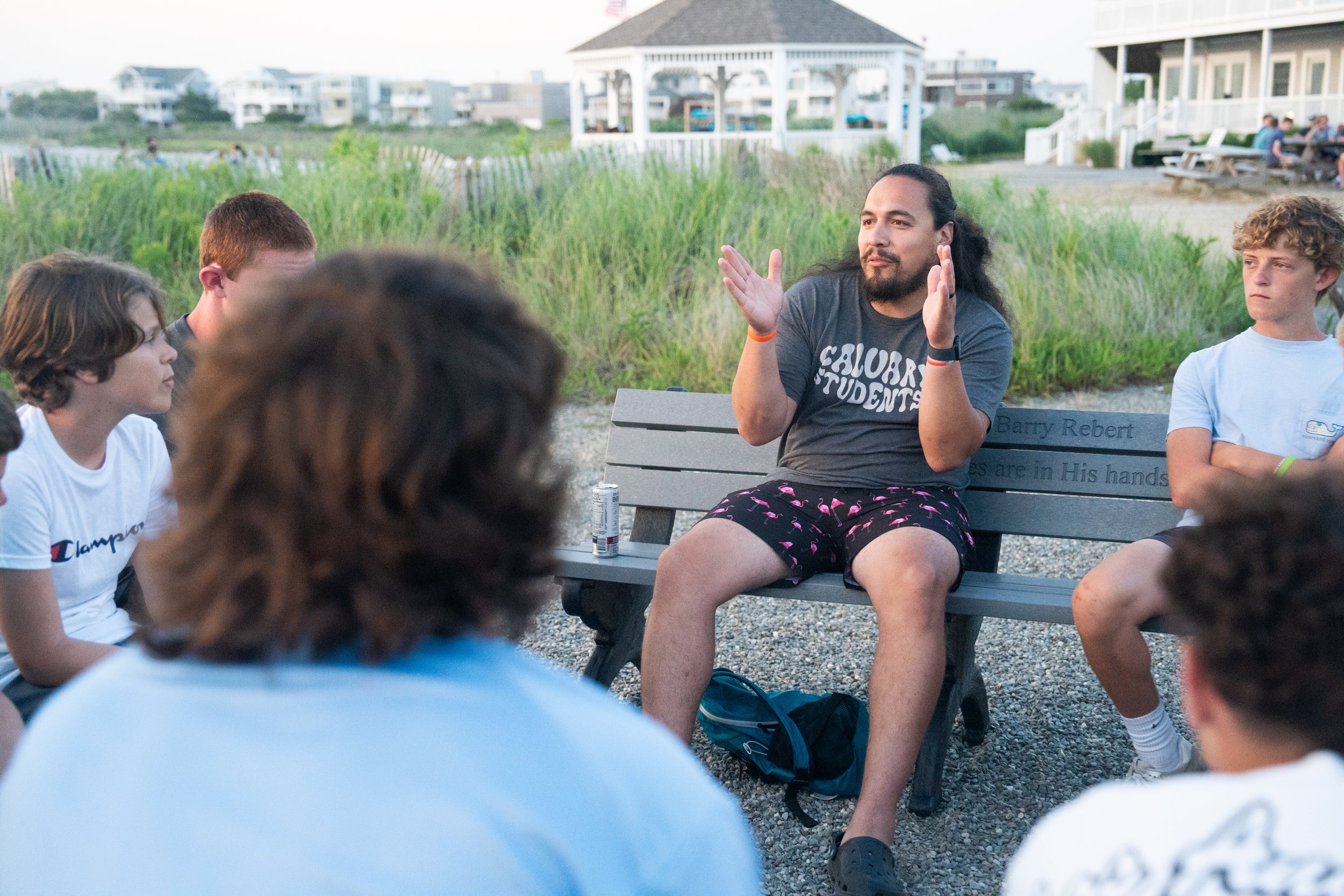 A man with long hair, wearing a dark shirt and shorts, sitting on a bench and talking to a group of young people outdoors by the beach, surrounded by grass and buildings in the background.