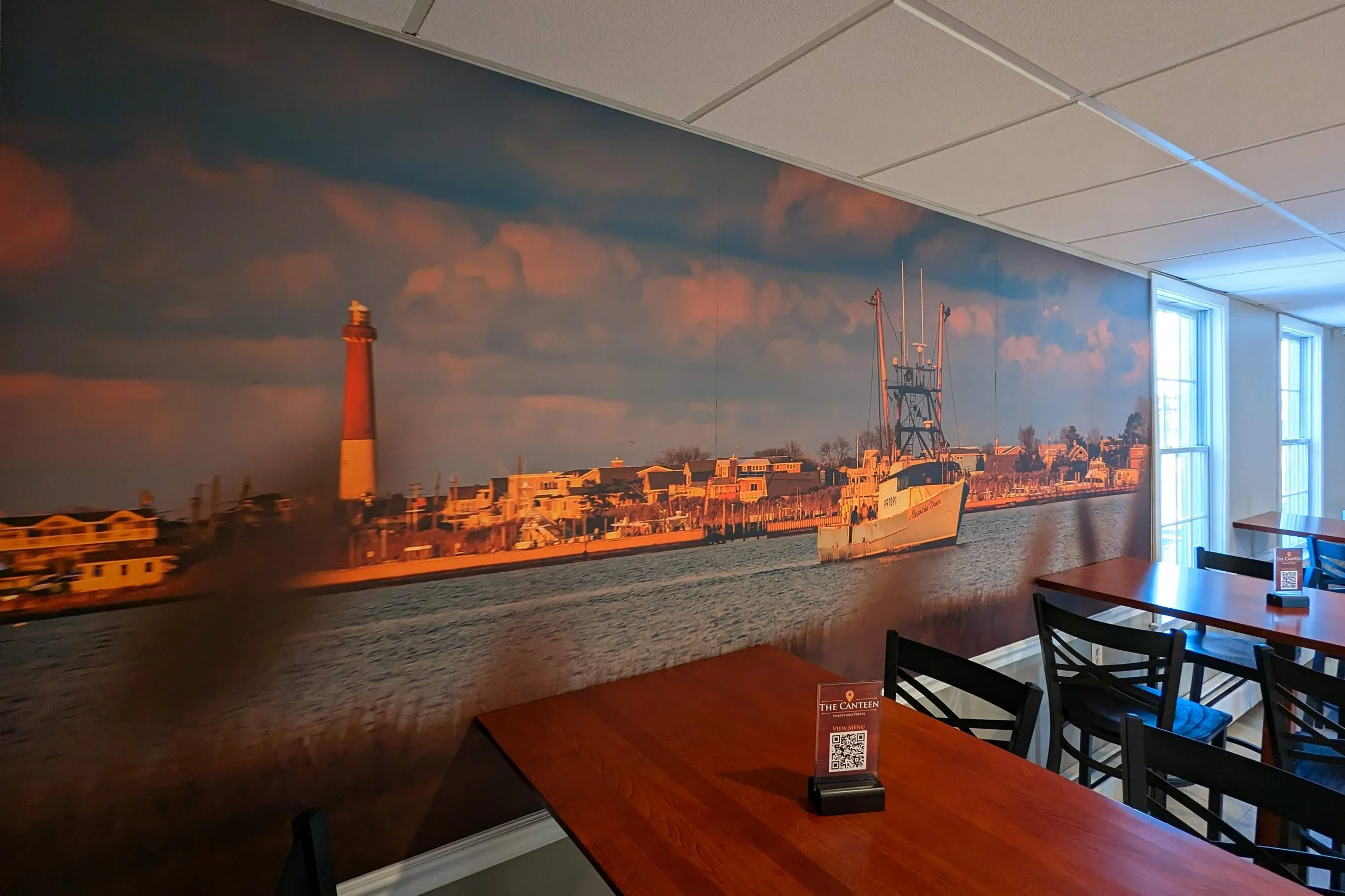 Large mural of a harbor scene with a lighthouse, boats, and waterfront buildings during sunset inside a restaurant with wooden tables and black chairs.