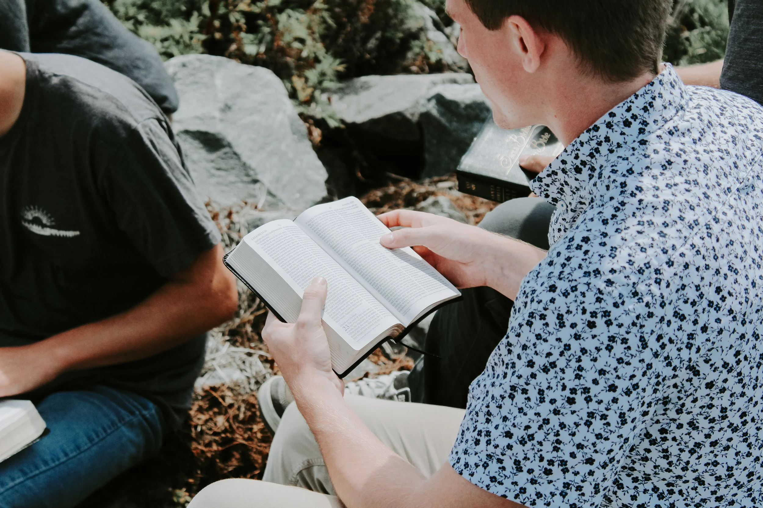 Two individuals are sitting outdoors, engaging with books. One person is holding open a Bible, and the other is reading a book with a floral pattern shirt. They are surrounded by rocks and nature.