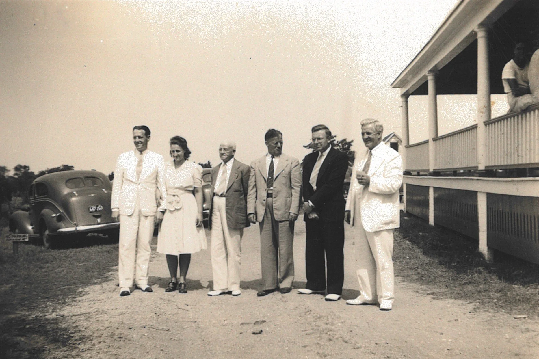 Black and white photo of six people standing outdoors near a vintage car and a building with porch, outdoors on a sunny day.