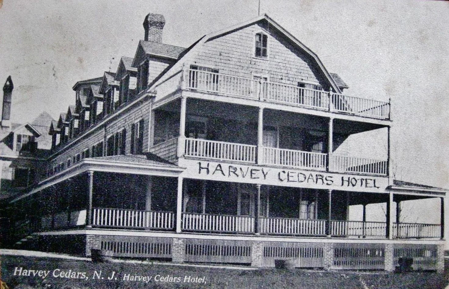 Black and white photograph of the Harvey Cedars Hotel building with three stories, wooden balconies, and a sign that reads 'Harvey Cedars Hotel' on the front. The image has a caption at the bottom indicating it is in Harvey Cedars, New Jersey.