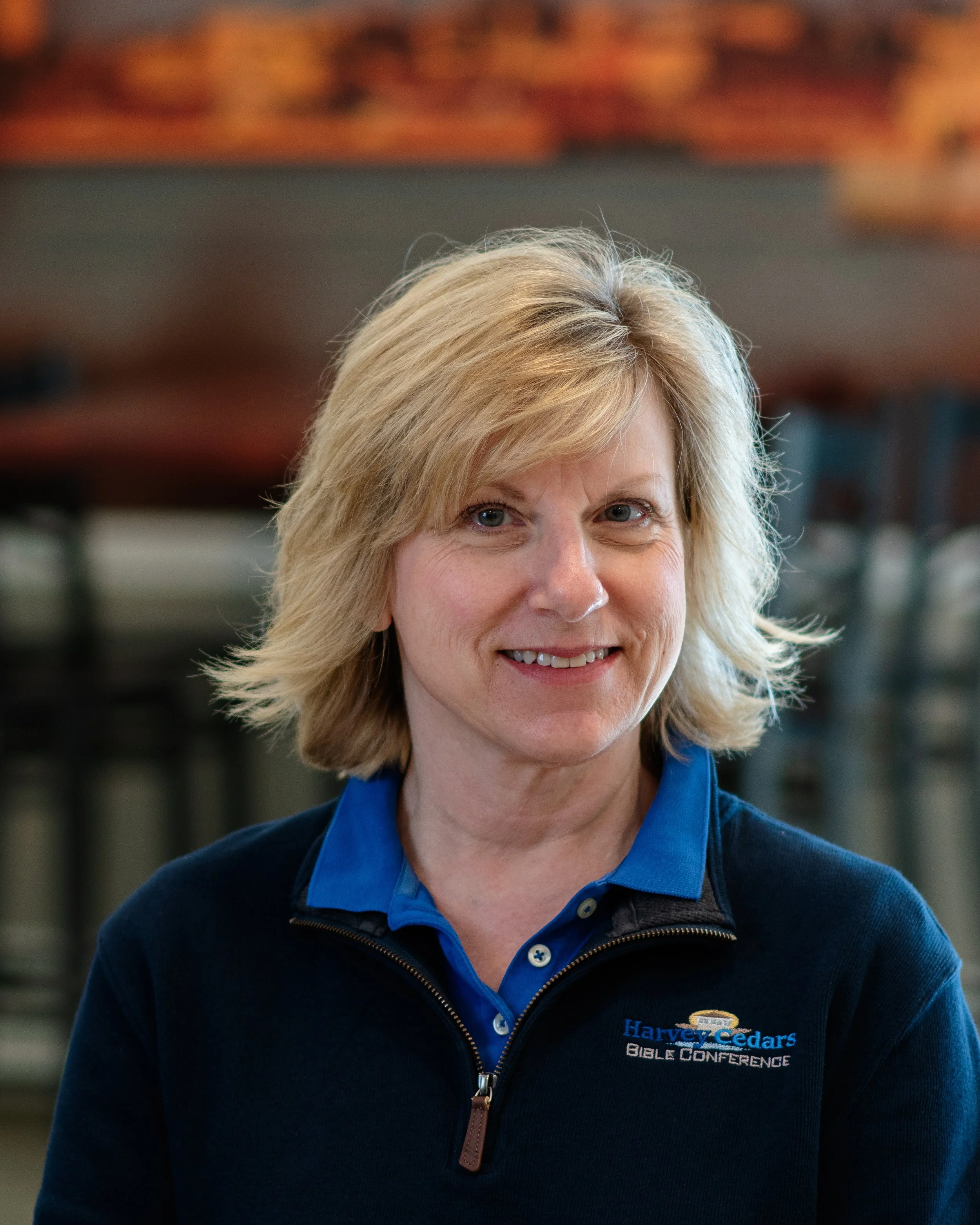 A smiling middle-aged woman with blonde hair wearing a Harvey Cedars Bible Conference jacket, indoors with blurred chairs and a painting in the background.