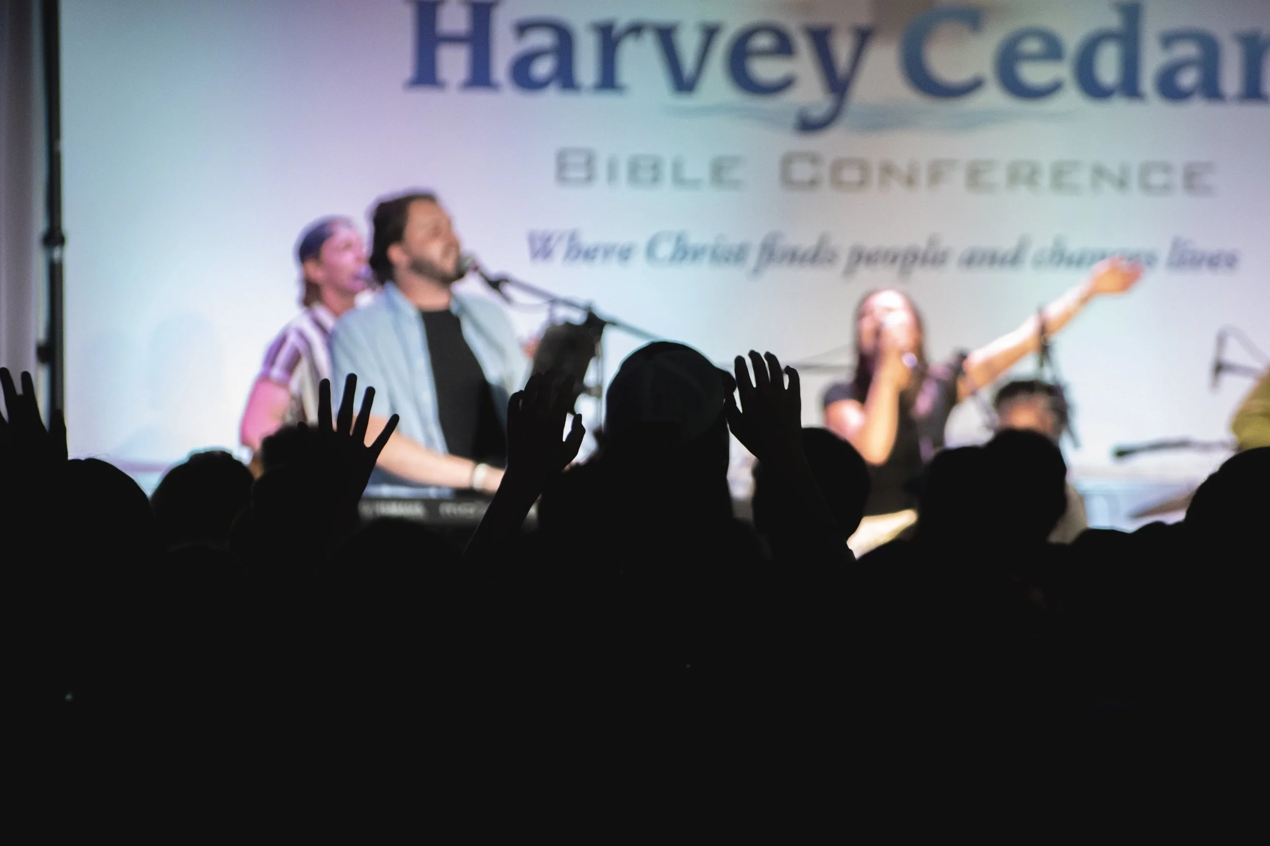Silhouettes of audience members with hands raised facing a blurred stage where a music group is performing during a Bible conference.