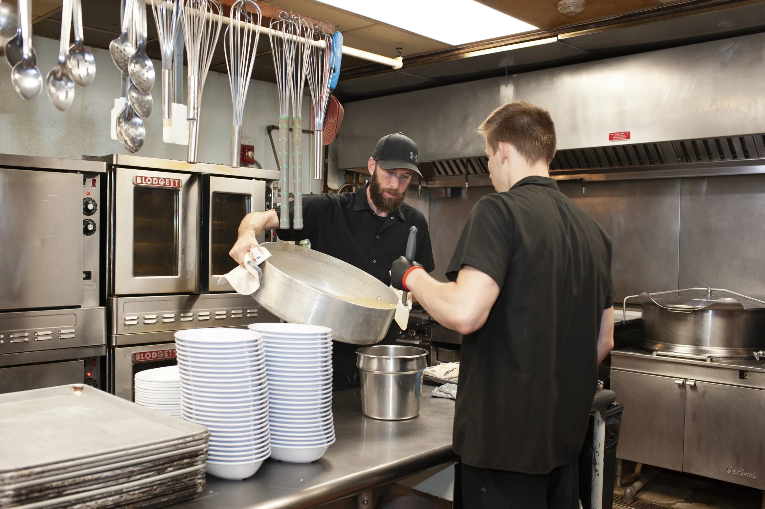 Two chefs in a commercial kitchen preparing food, with one pouring soup into a bowl while the other assists, surrounded by kitchen equipment and stacked bowls.