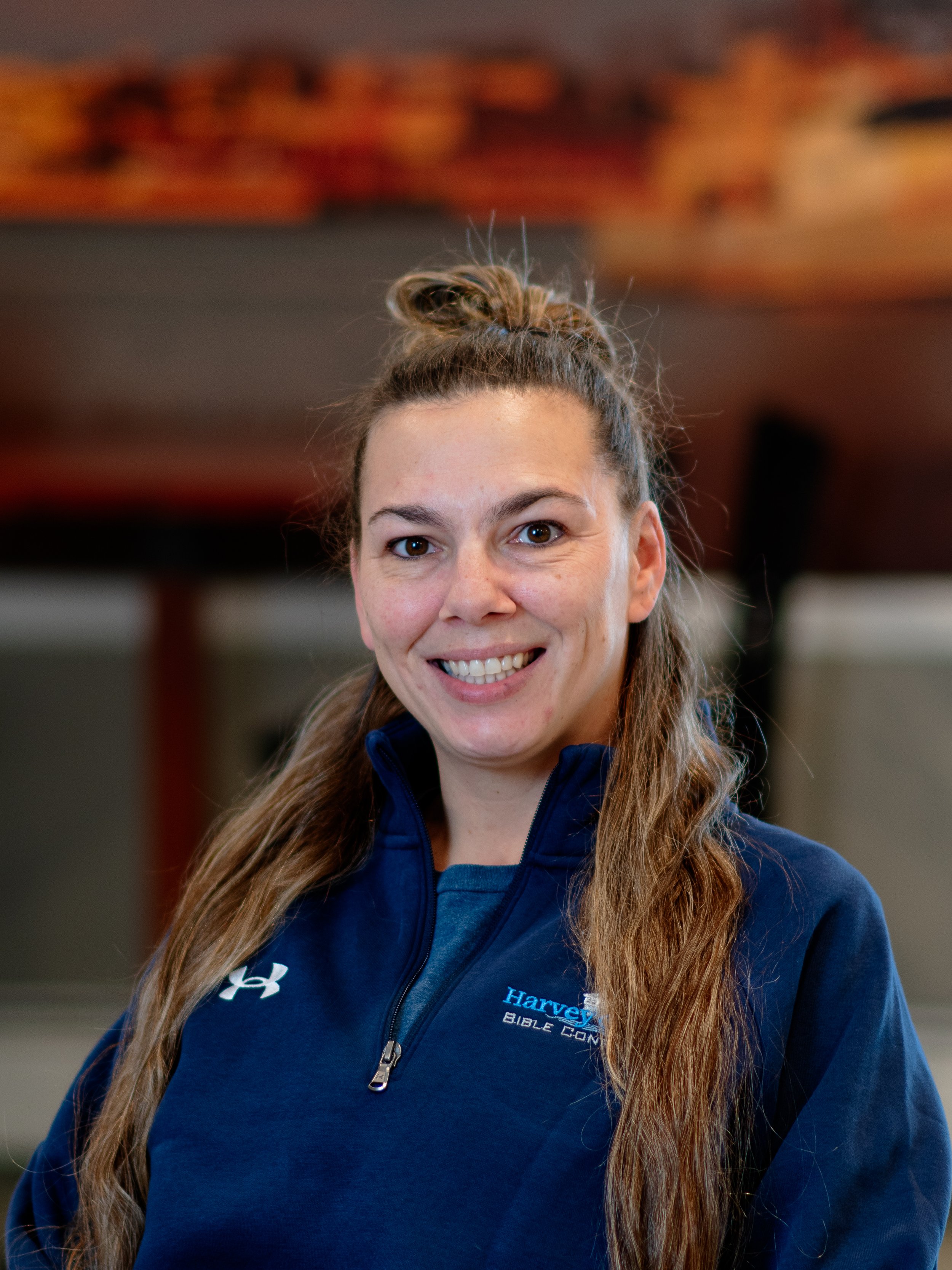 Close-up of a smiling woman with long brown hair tied in a bun, wearing a blue hoodie with 'Harvey Bible Camp' embroidered on it, indoors with a blurred background.