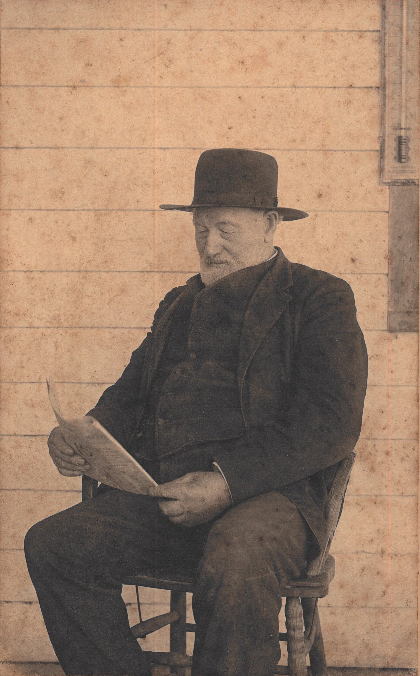 An elderly man with a beard and wearing a large black hat sits on a wooden chair, reading a newspaper in front of a rustic wooden wall.