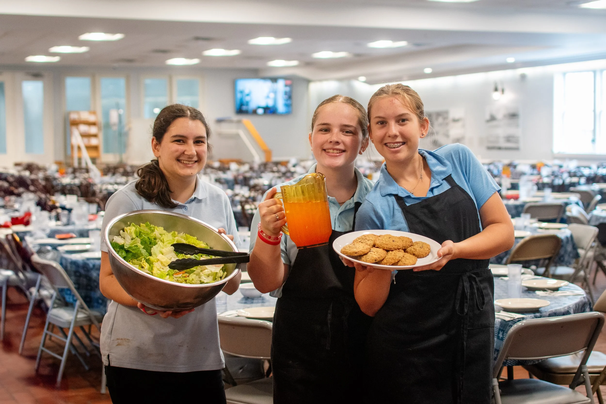 Three young girls in a cafeteria holding salad, iced tea, and cookies.