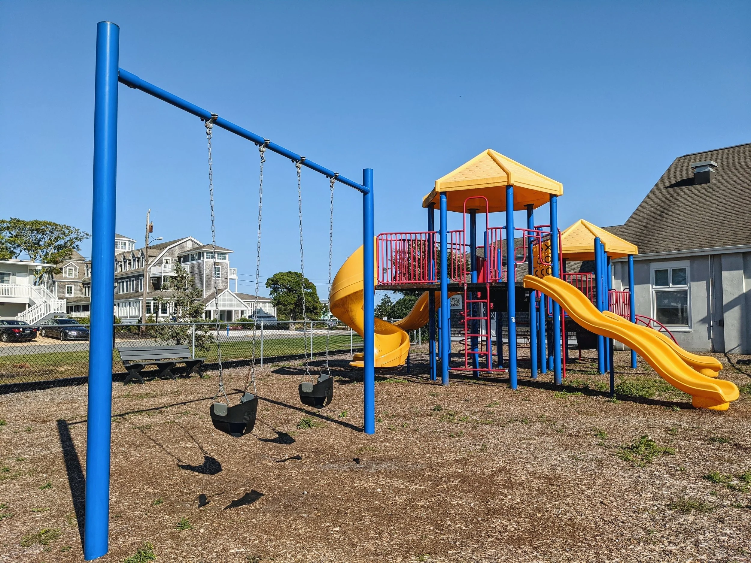 Colorful playground with yellow slides, red railings, blue support posts, and black rubber swings on a dirt ground, with residential houses and a chain-link fence in the background.