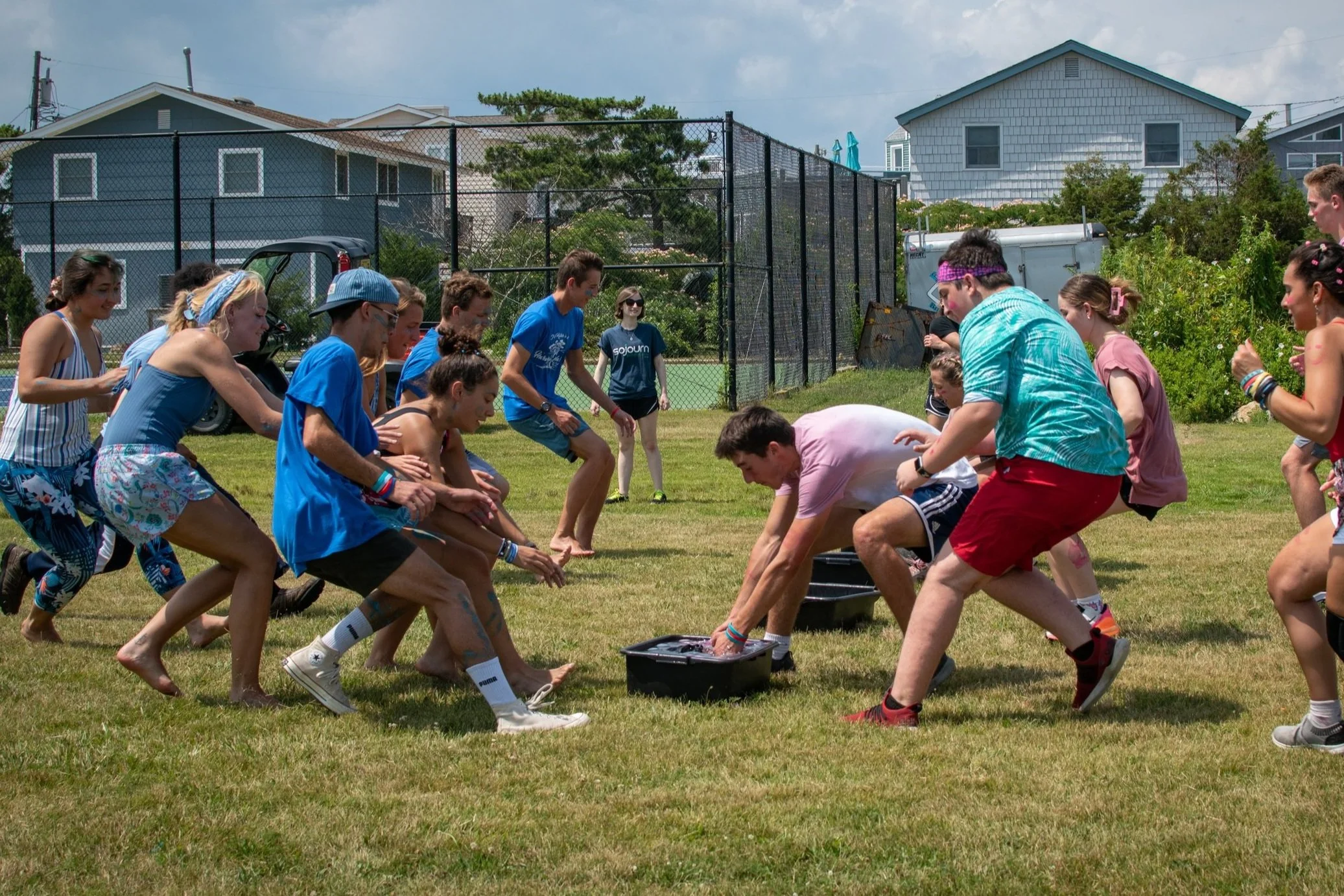 People playing a game of tug of war on a grassy field with houses and a tennis court fence in the background.