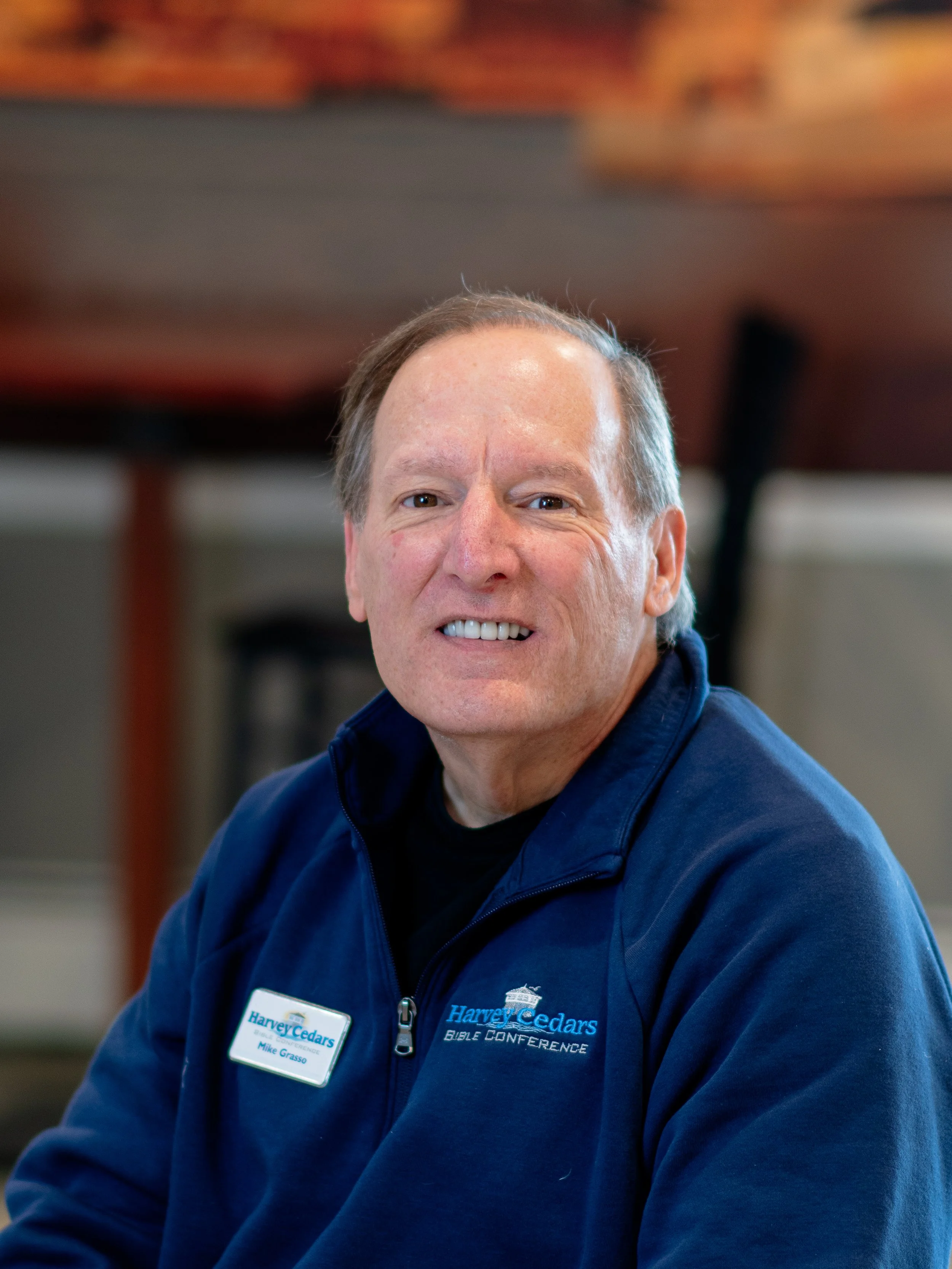 A smiling man wearing a dark blue jacket with a name tag and a logo that reads 'Harvey Cedars Bible Conference.'