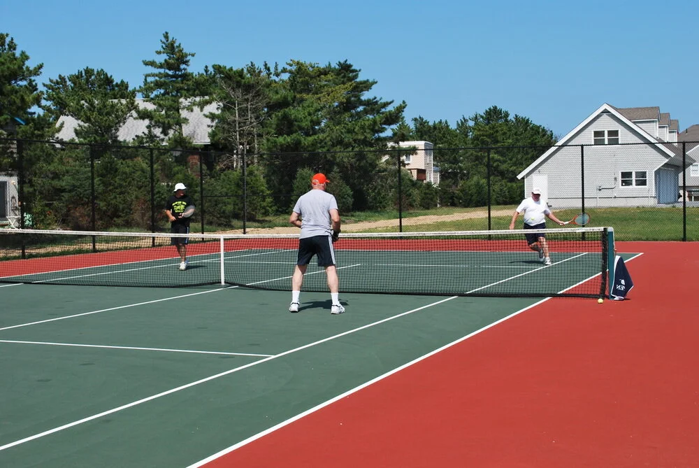 Three men playing pickleball on an outdoor court surrounded by trees and houses under a clear blue sky.