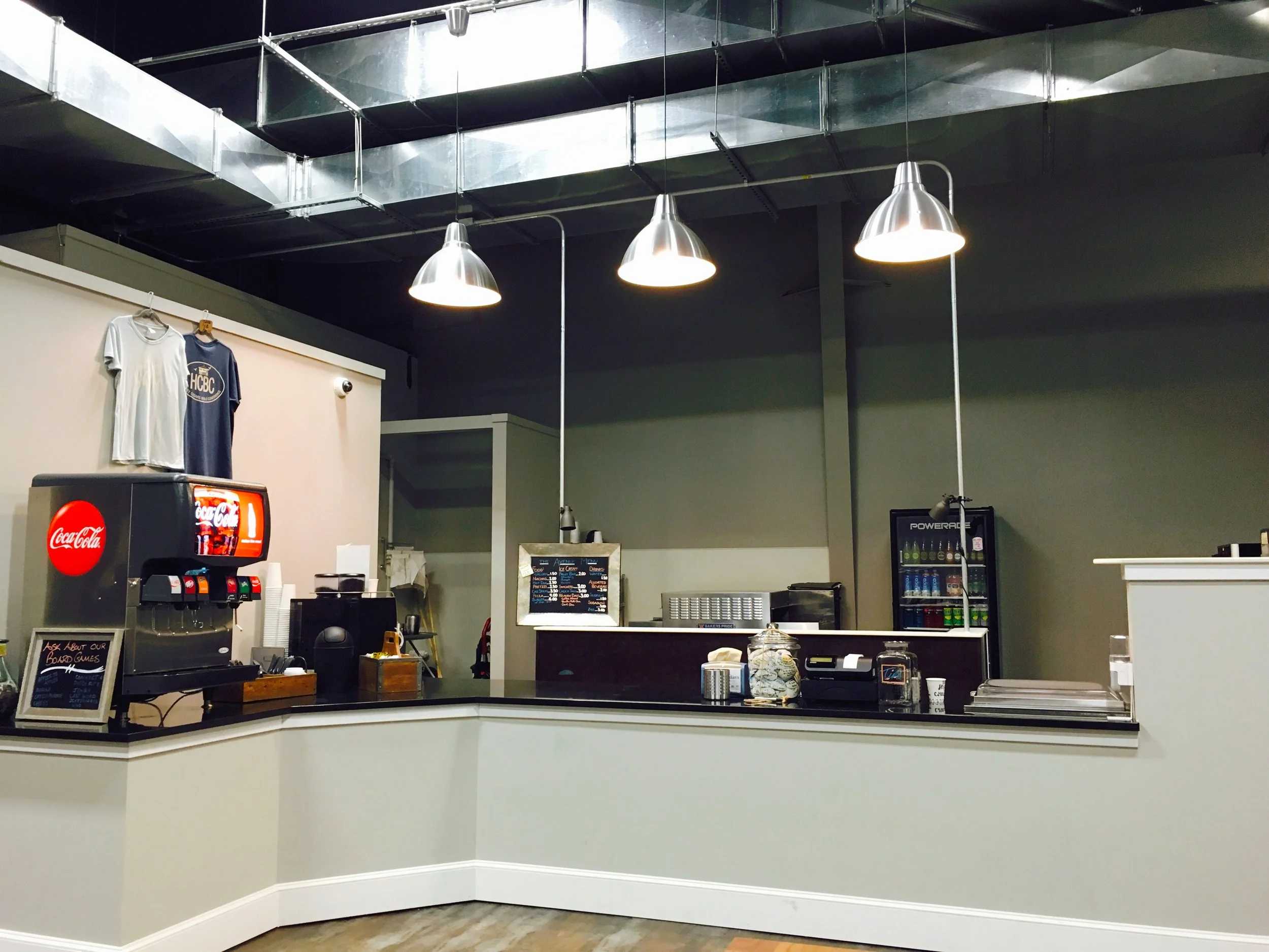 Empty cafe counter with a Coca-Cola soda fountain, a small fridge, and hanging pendant lights in an industrial-style interior.