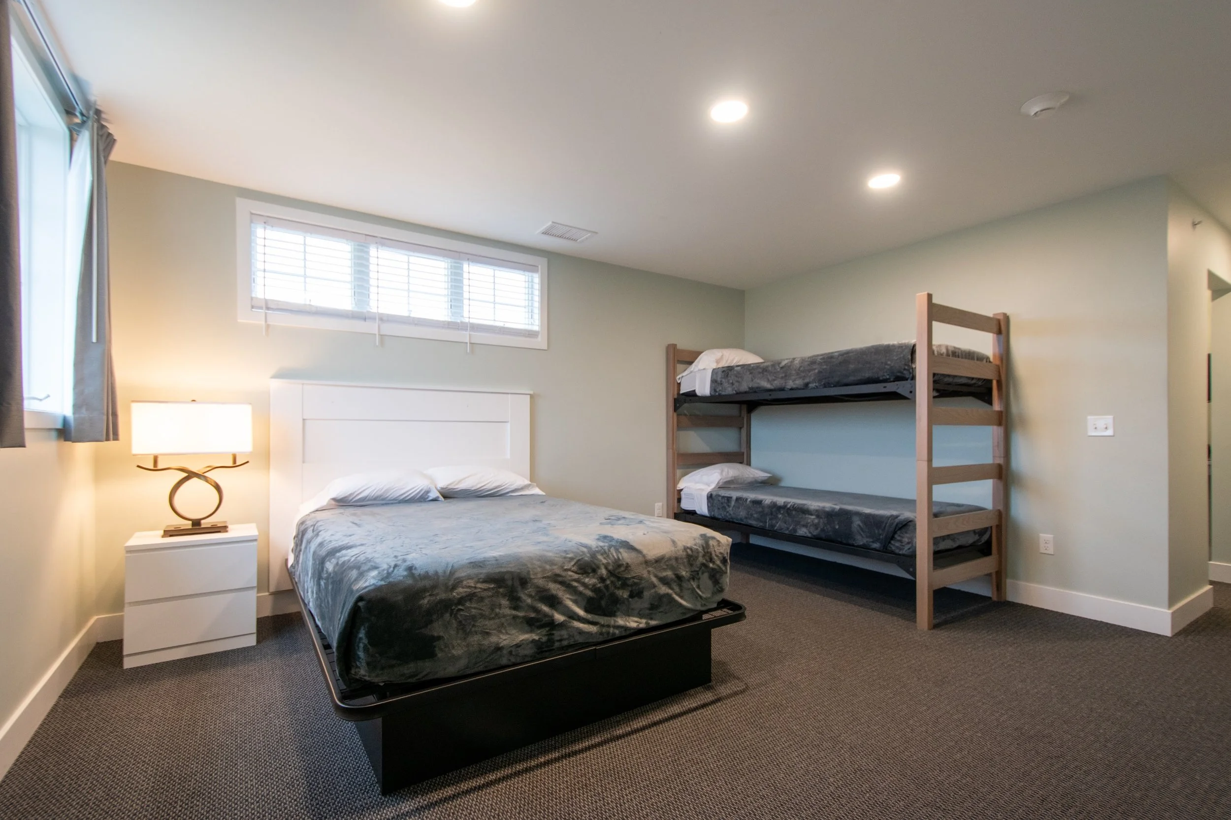 A bedroom with a queen-sized bed, a nightstand with a lamp, and a window with blinds. There are two twin beds stacked on a wooden frame in the corner. The room has beige walls and a carpeted floor.