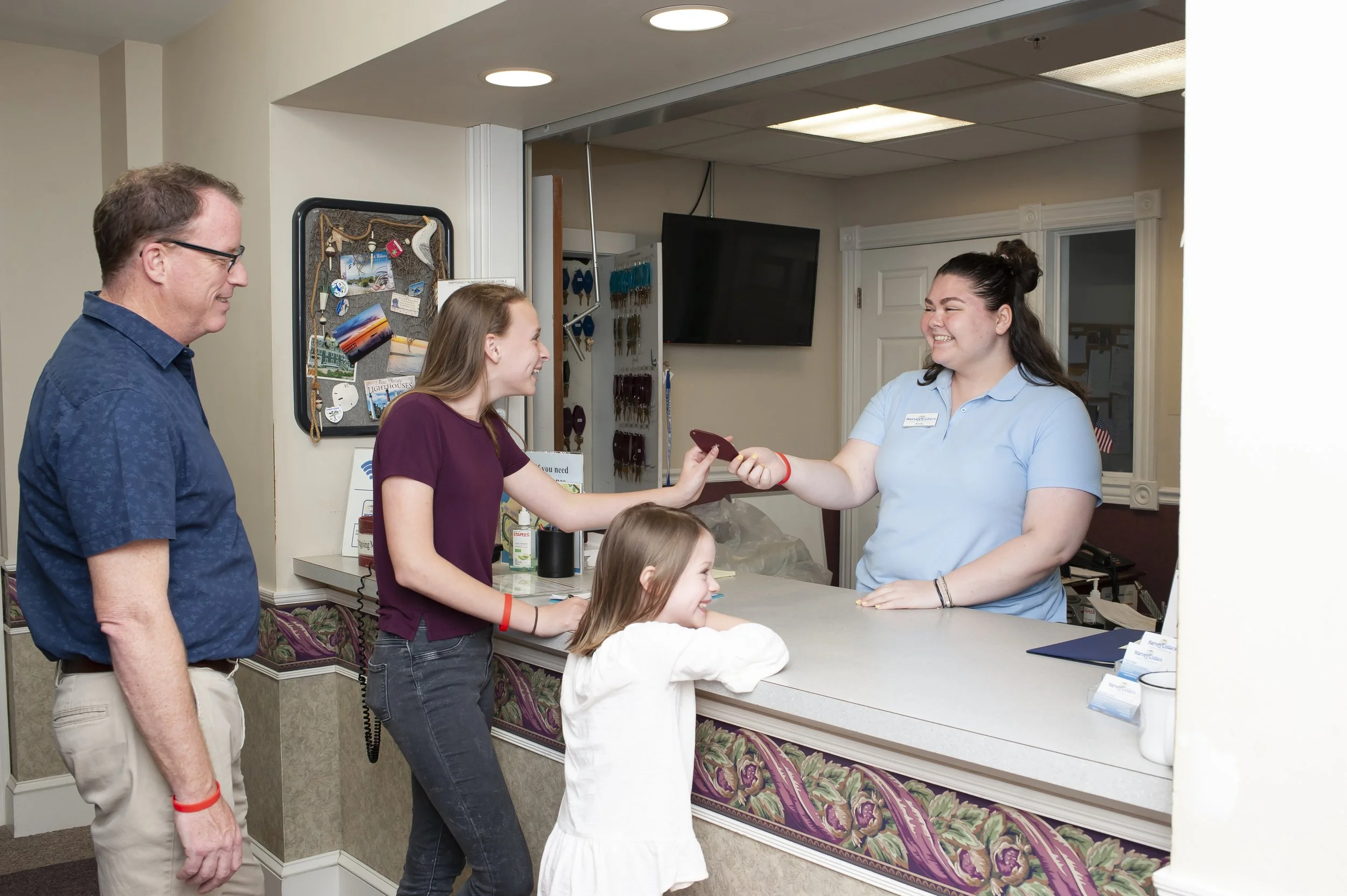 A young girl hands a credit card to a cashier at a reception desk, with a man and another girl standing nearby, smiling in a cheerful indoor setting.