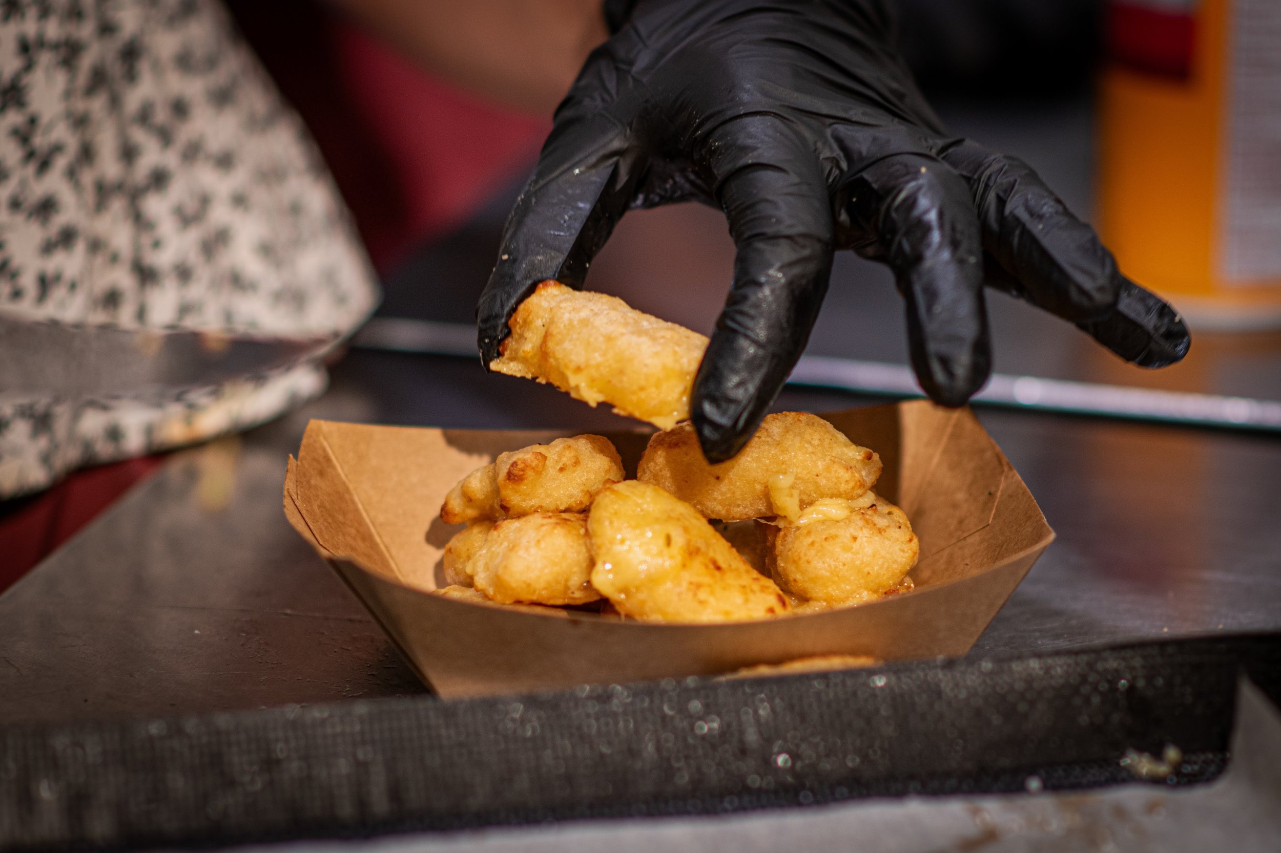 A person wearing a black glove is picking up a piece of fried food from a paper tray filled with golden-brown fried nuggets on a black tray.